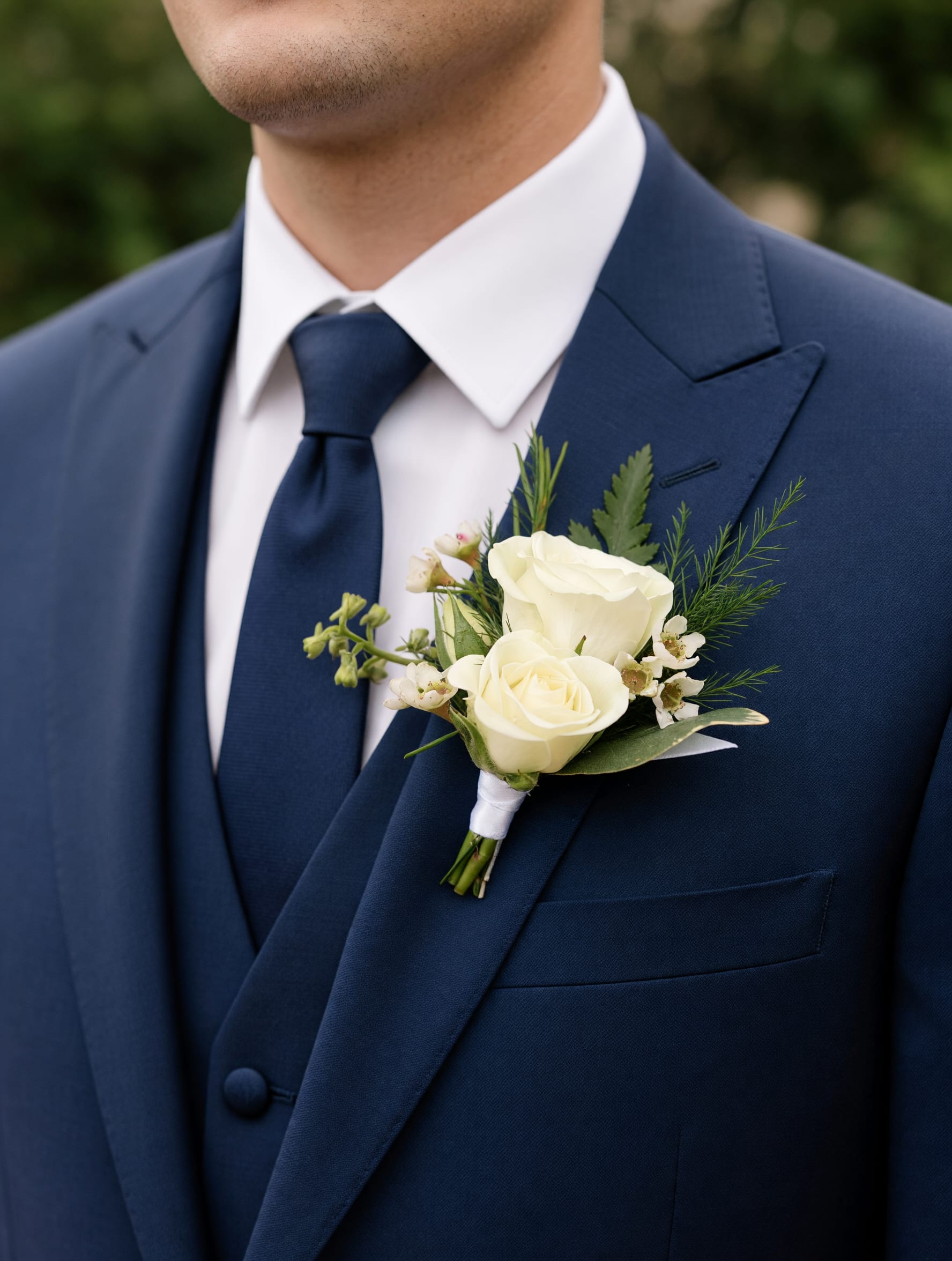 Boutonniere - Classic White - The Classic White Boutonniere consists primarily of white spray roses, with a pop of complimentary filler flower and greenery. Designed in a neutral color palette, this piece beautifully complements any outfit without overpowering it, allowing your attire to remain the focal point while adding a petite floral accent.  Select the deluxe option to order a bout. &amp; corsage set!