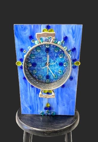 #A137 - Blue Mantle Clock with trivet - A sculptural blue clock that reads like modern art - perfect as a housewarming gift or a thoughtful birthday present for someone with eclectic taste. This decorative clock artwork is presented as a mantle-style decoration (with coordinating trivet) that doubles as home décor, making it a unique gift for anniversaries, new homes, or to mark a special occasion in a stylish interior.   The piece features layered shades of blue on a painted panel and tall rectangular body, punctuated by a large circular metal clock face. Clustered glass beads in cobalt, aqua, yellow, and hints of purple and white create a mosaic-like, textured surface that catches light and invites closer inspection. Beaded embellishments and an art-panel aesthetic give it a handcrafted, contemporary vibe rather than a clean modern look. Give this to the person who loves bold color and modern design - it's the kind of present that makes someone feel seen for their style. The durable wooden base and metal clock face make it display-ready on a mantel, shelf, or console; no floral care required, just choose the spot where the blue tones and glass beads will pop.