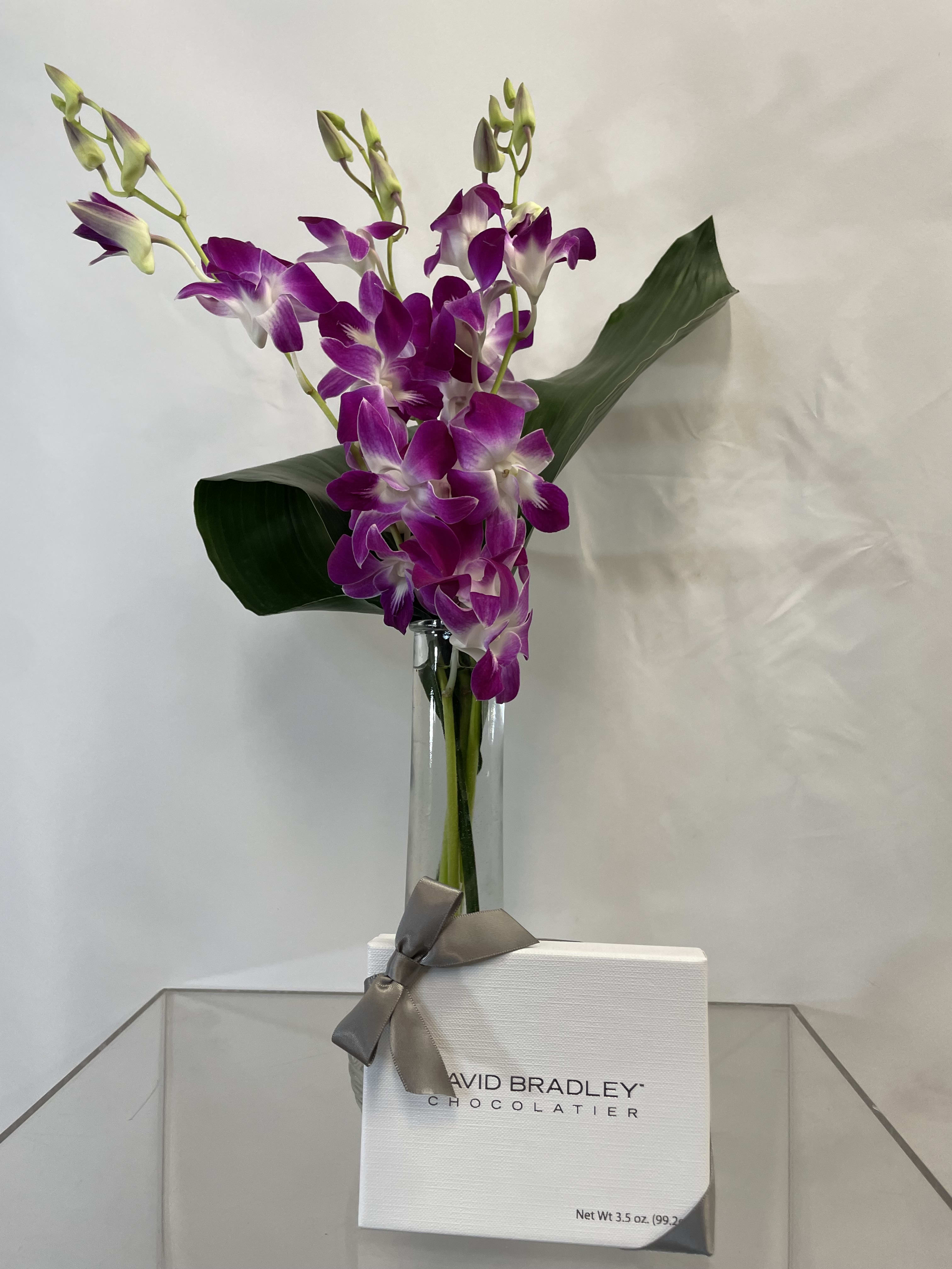 Purple orchids in a glass vase with a white chocolate box tied by a gray ribbon.
