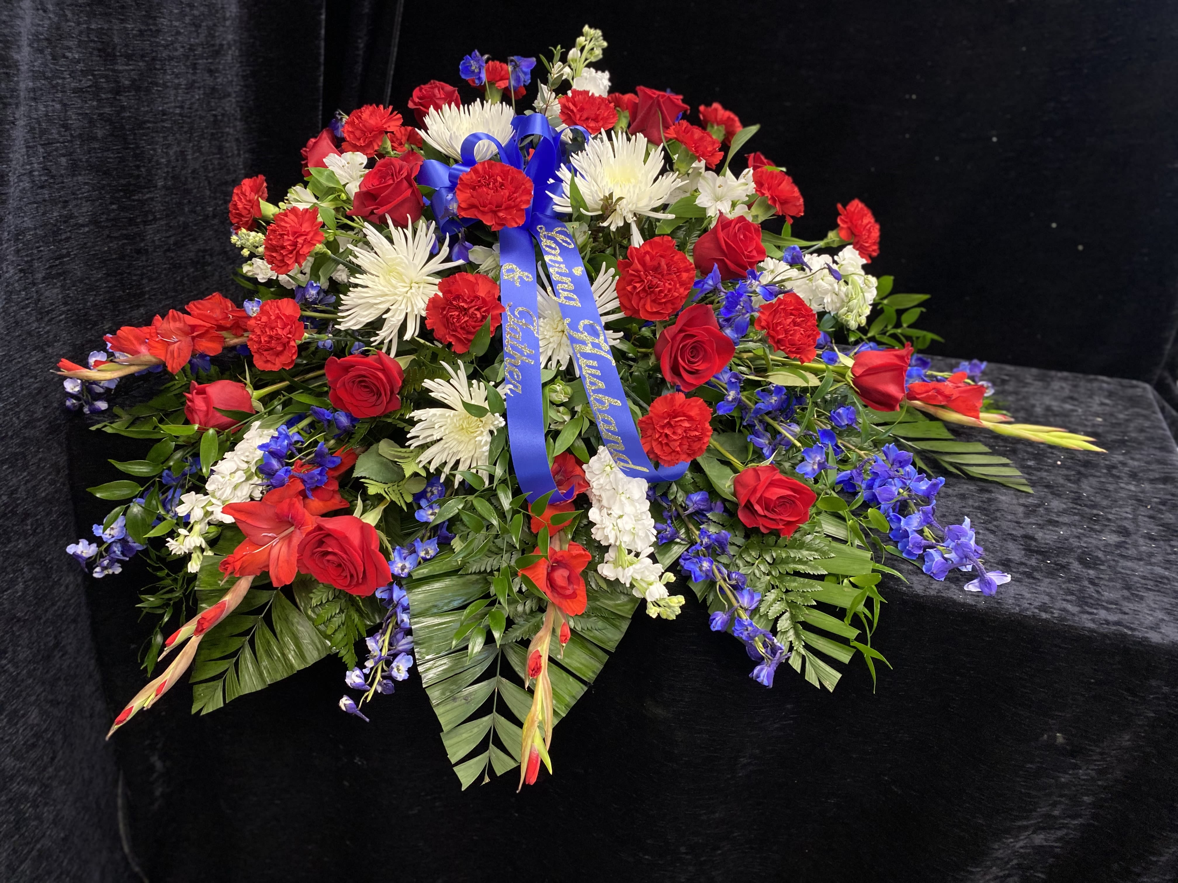 Memorial Casket Spray - A broad, low casket spray crafted in a bold red, white, and blue palette to honor a life with dignity and patriotic grace. This memorial casket spray is arranged for funerals and commemorative services, the kind of floral tribute that makes the person you're remembering feel celebrated and respected. The strong combination of red roses and carnations with white spider chrysanthemums and blue delphinium creates an immediate visual statement suited for military, first-responder, or national memorial occasions.  Appropriate for funerals, memorials, or patriotic remembrances, this arrangement communicates honor and heartfelt sympathy to the family and attendees. Arrives ready to place; minimal care is needed beyond keeping the foam hydrated if the service is delayed. If you need help coordinating delivery to a funeral home or service location, we can assist to ensure the spray arrives on time and in proper condition.