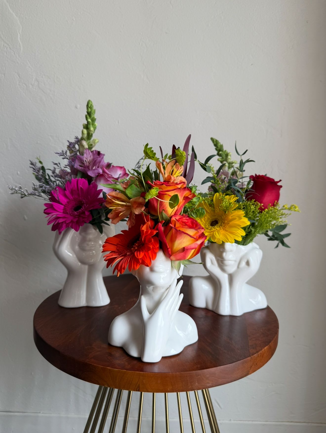 Three colorful flower arrangements in white face-shaped vases on a round table