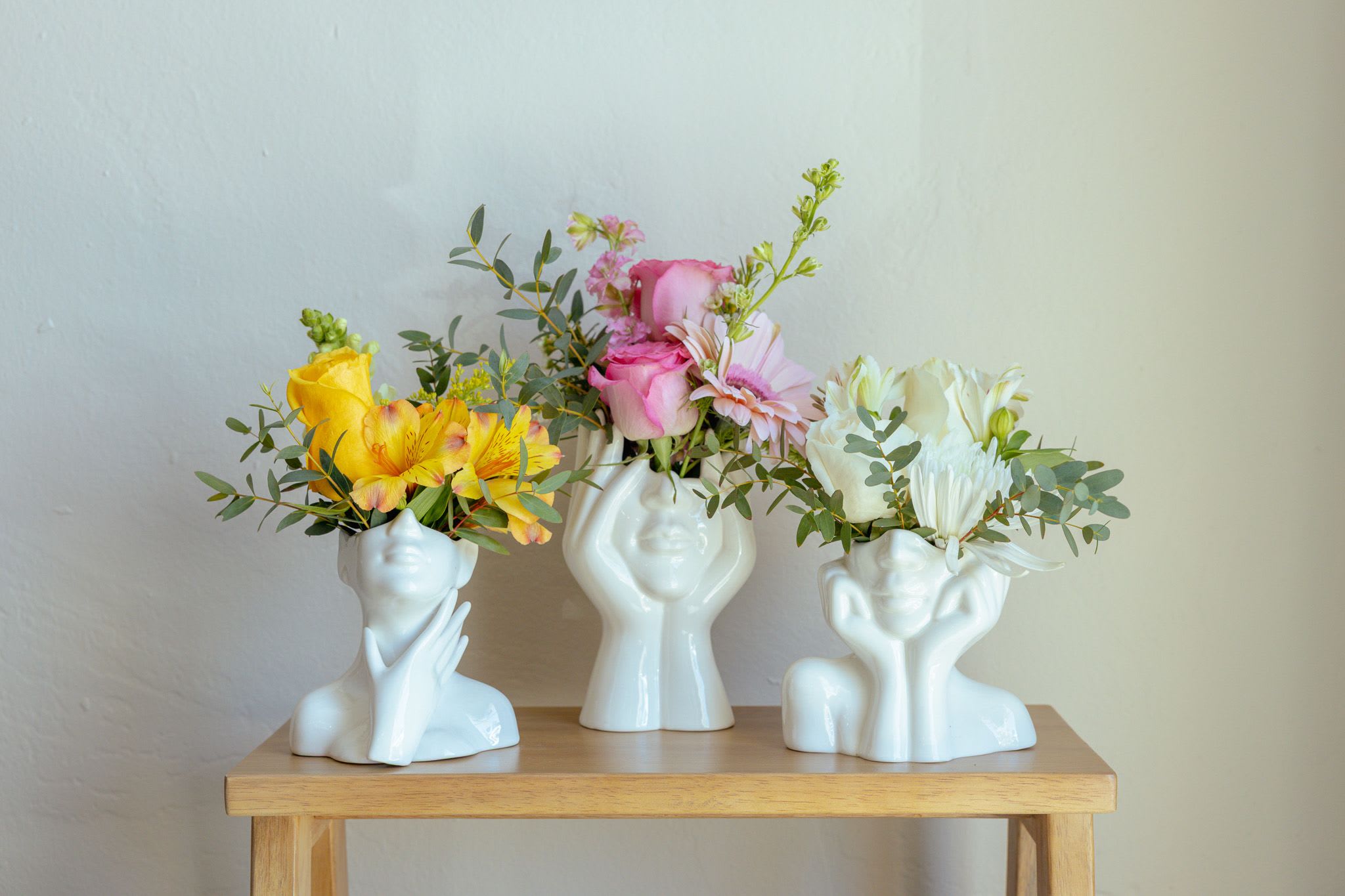 Three small floral arrangements in white face-shaped vases on a wooden table