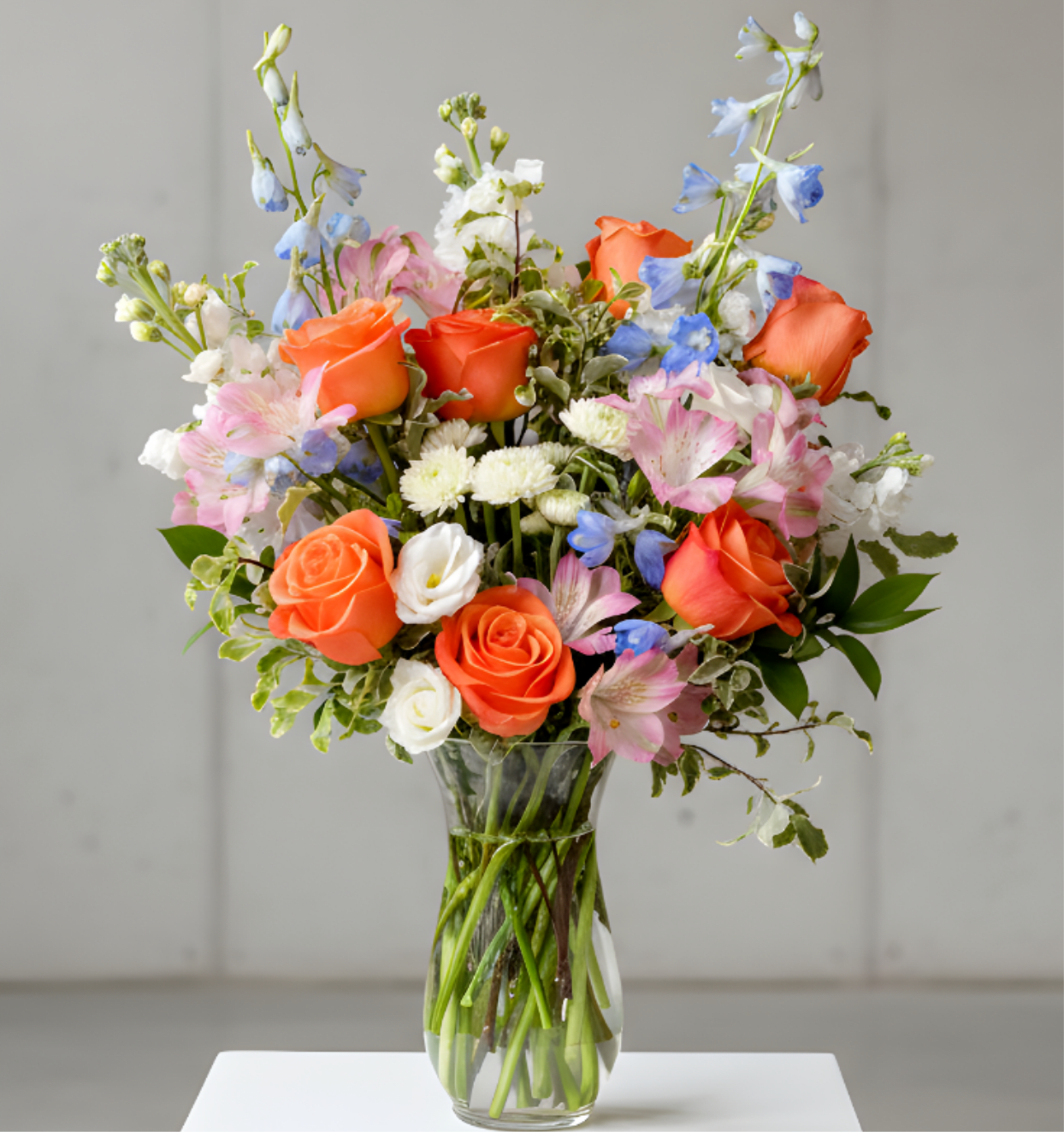 Tall mixed arrangement of orange roses, pink blooms, and blue delphinium in a clear glass vase