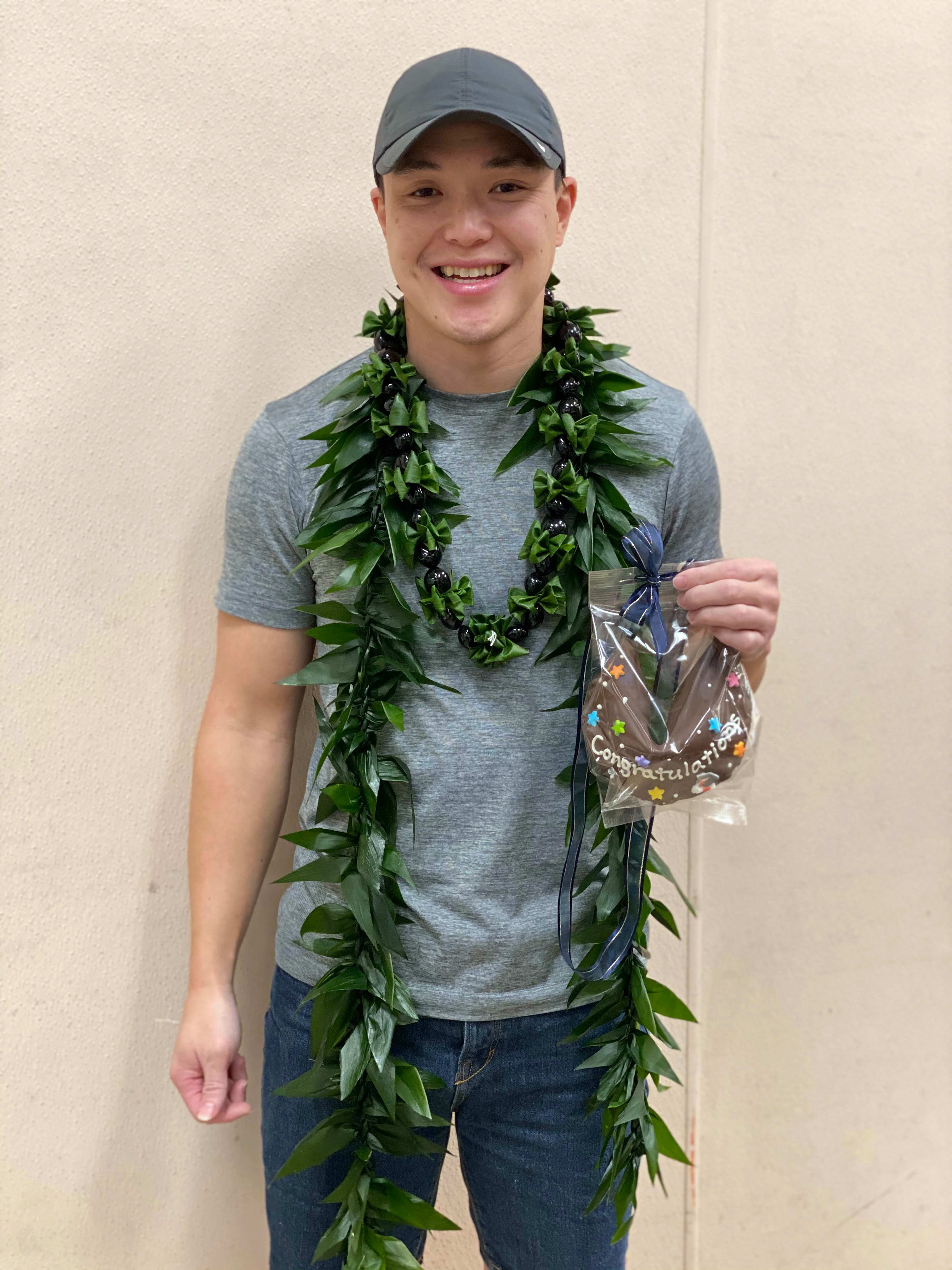 Person wearing a long green leaf lei and holding a small wrapped congratulations gift.