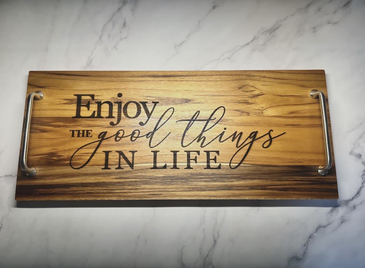 “Enjoy the Good Things in Life” Wooden Serving Tray - Bring warmth and timeless style into the home with this beautifully crafted teak wood serving tray. Made from durable teak, known for its rich grain and long-lasting quality, this piece offers both function and elevated style. The message, “Enjoy the good things in life,” is carefully burned/etched into the wood, giving it a natural, rustic character that won’t fade over time.  Finished with sturdy metal handles for easy carrying, this tray is perfect for serving drinks, breakfast in bed, or styling on a coffee table, ottoman, or kitchen island. Perfect gift for newlyweds, anniversary, new home owners, or birthday's!  Details:  Dimensions: 21.5" x 9" Material: Teak wood with metal handles Finish: Natural teak wood with burned/etched lettering Style: Rustic, coastal, modern farmhouse  A beautiful piece for everyday use or gifting—because the little moments are the ones worth enjoying most.