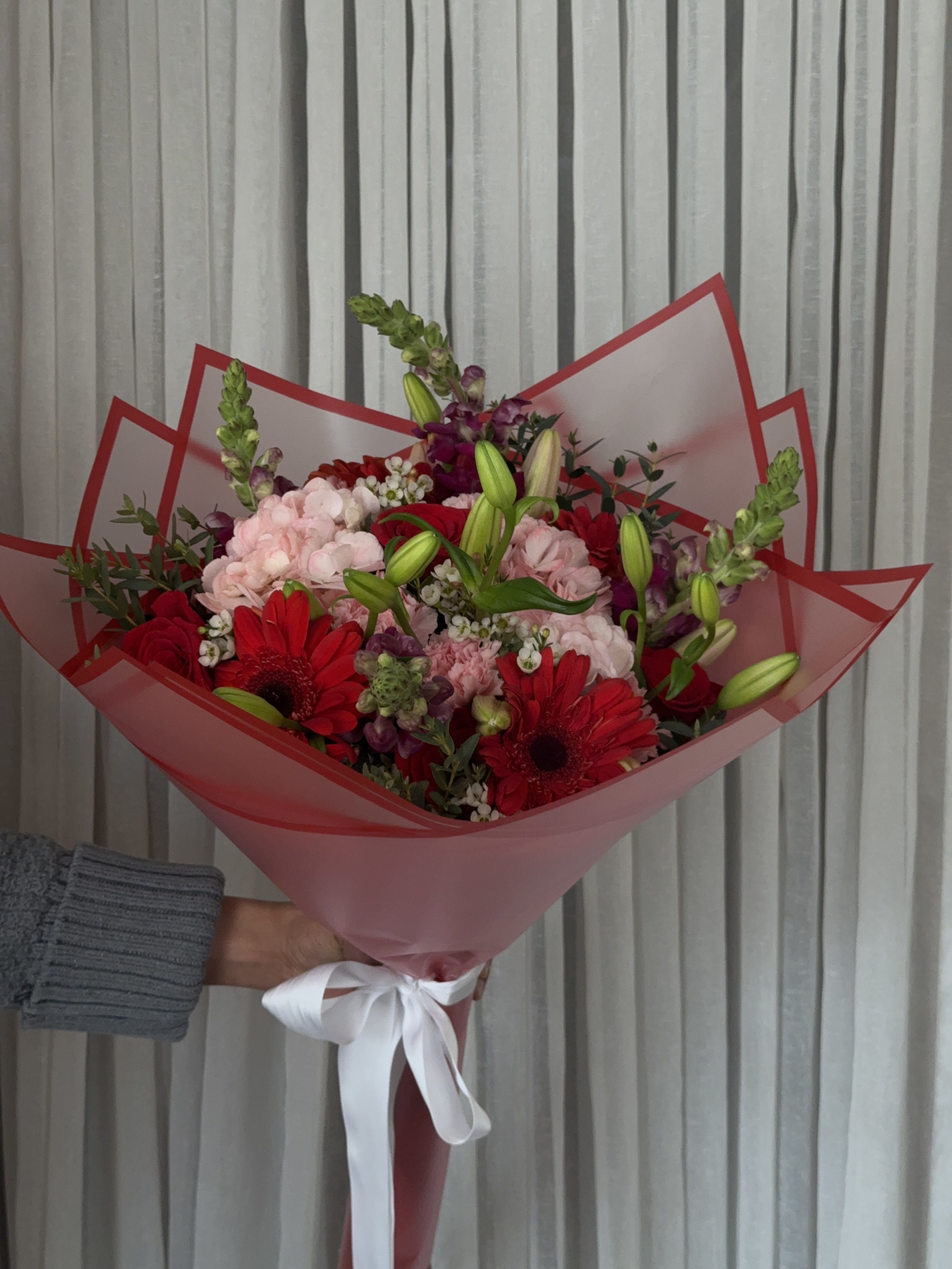 Handheld bouquet of red and pink flowers wrapped in pink paper with a white ribbon