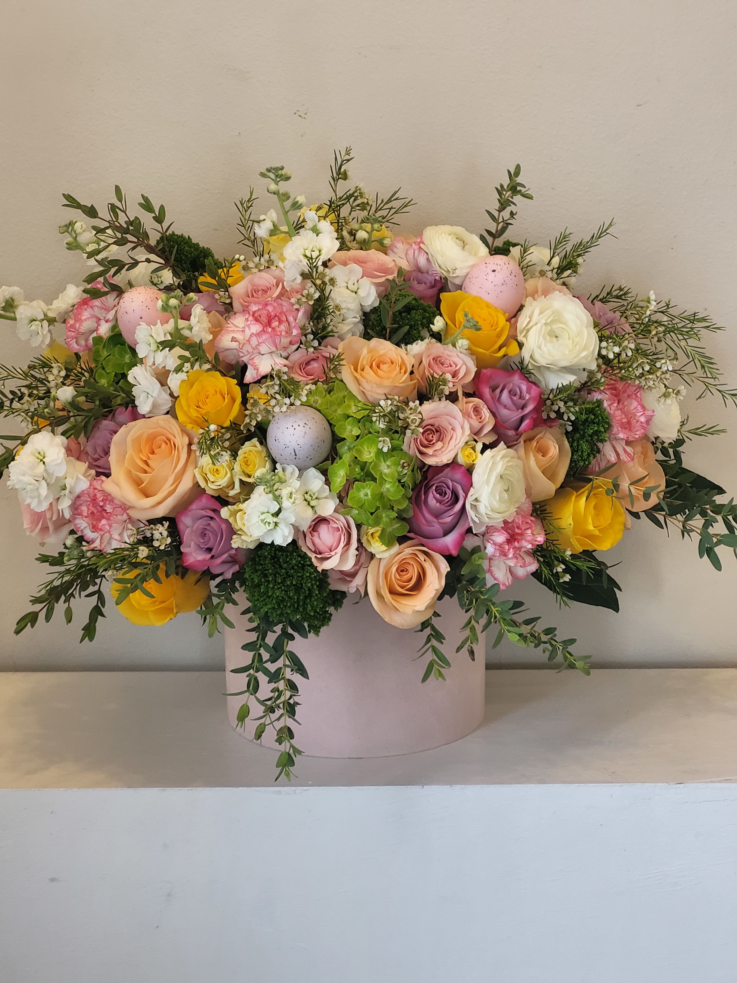 Spring Pastel Everyday Bouquet - A lush, garden-style spring bouquet arranged low and full in a round pink hat box-perfect as an Easter centerpiece or a cheerful spring gift. This flower arrangement blends roses, carnations, ranunculus, stock, and hydrangea in a soft pastel palette of blush, peach, cream, lavender and bright yellow so the person you're thinking of will feel celebrated and seen.   The composition is dense and rounded with airy branching greenery for a natural, garden-texture that reads abundant without being fussy. Compact clusters of green hydrangea balance the softer roses and ruffled carnations, while white ranunculus and small accent blooms brighten the design. The flowers arrive nestled in a pale pink cylindrical hat box (or white container option), making it an easy display piece that requires no vase.  Ideal for Easter, a spring birthday, or a "just because" moment, this arrangement suits anyone who loves romantic pastels and a joyful, seasonal statement. Care is simple-keep the floral foam hydrated and out of direct sunlight to maintain freshness-perfect for a lasting gift that keeps the recipient smiling.