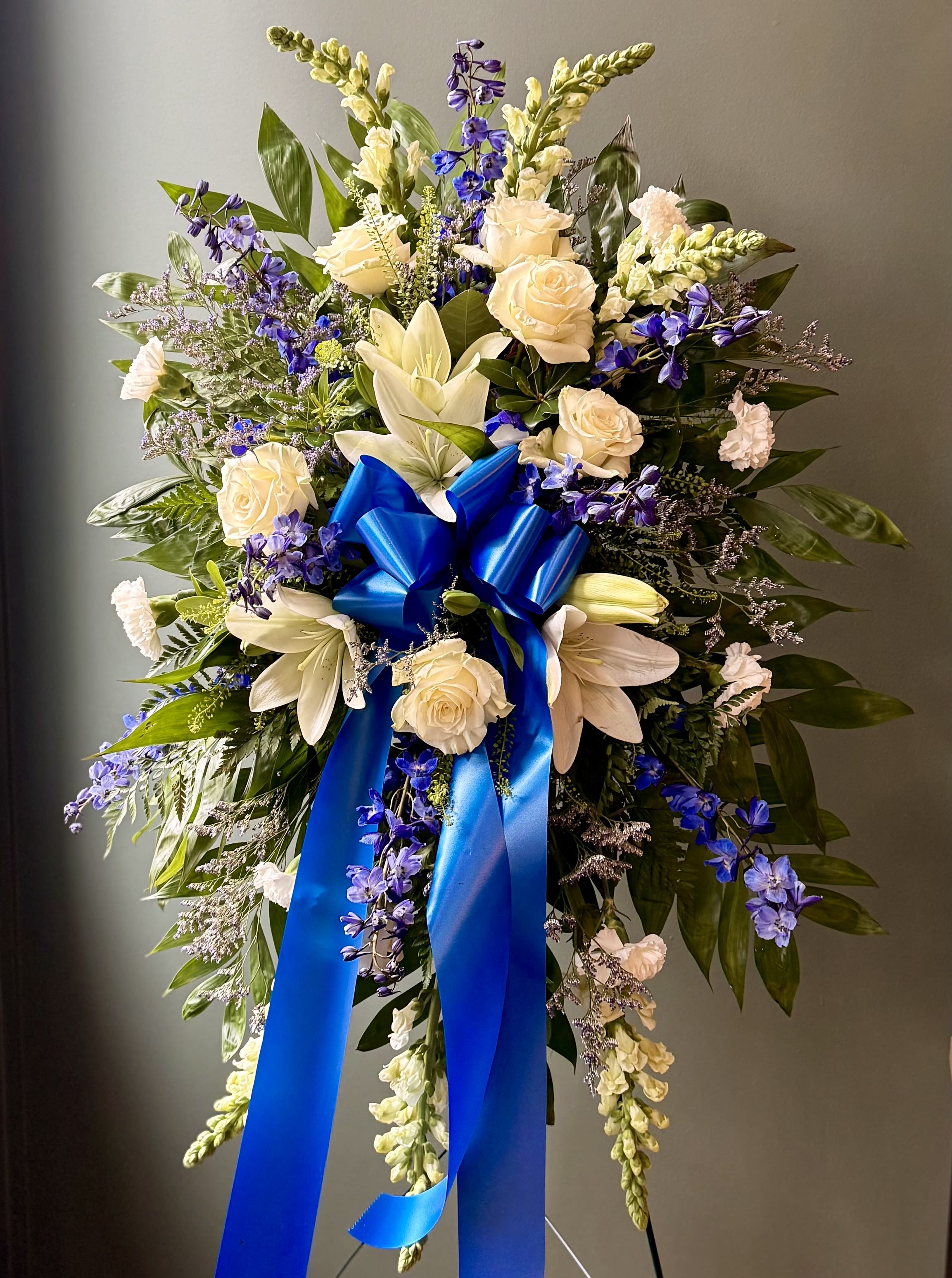 Standing spray of white roses and lilies with blue flowers and a large blue bow on an easel.