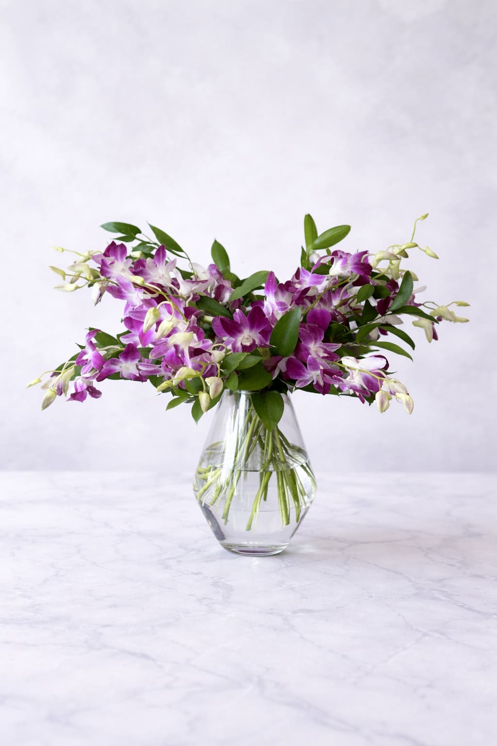 Orchid Bliss - A dramatic spray of purple-and-white dendrobium orchids makes a memorable birthday bouquet, anniversary gift, or wedding centerpiece. Orchid Bliss presents a wide, airy cluster of blooms that spill and cascade from a clear glass vase, giving the arrangement a fresh, slightly tropical elegance that reads celebratory and refined for the person you're honoring.