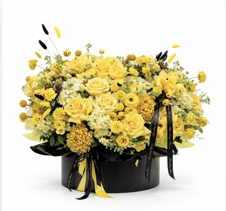 Alexis &amp; The Bees - Playful, radiant, and full of personality—Alexis &amp; The Bees is a sunshine-soaked arrangement designed to turn heads. Lush yellow roses, golden mums, and soft cream blooms come together in a rich, textural composition, accented with whimsical bee details that bring movement and charm.  Set in a sleek black vessel and finished with flowing ribbon, touches of faux bumblebees, this piece strikes the perfect balance between elevated and fun. It’s bright, a little unexpected, and effortlessly chic—made for anyone who loves a bold pop of color with a touch of sweetness.
