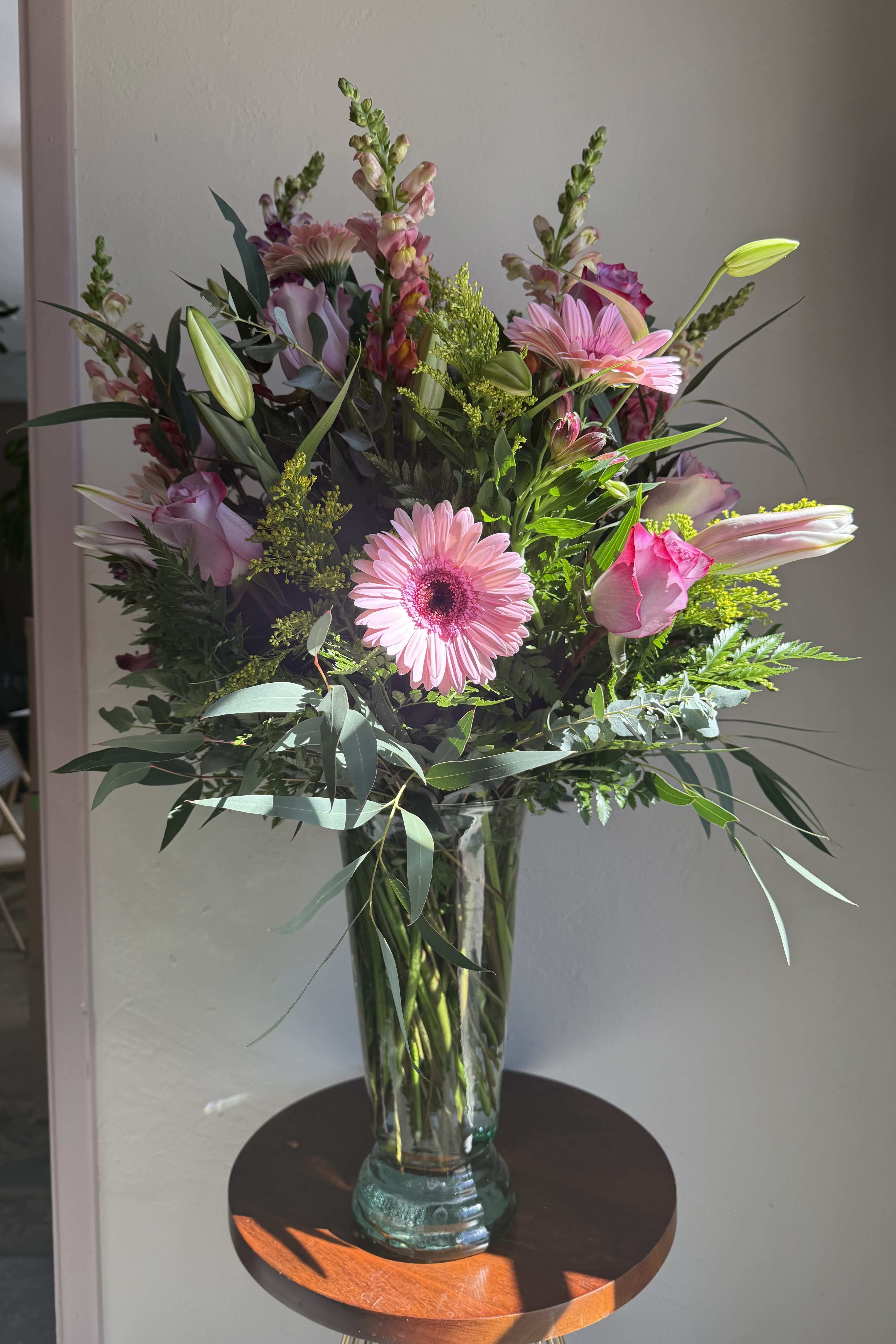 Tall arrangement of pink gerbera daisies, roses, lilies, and snapdragons in a clear glass vase