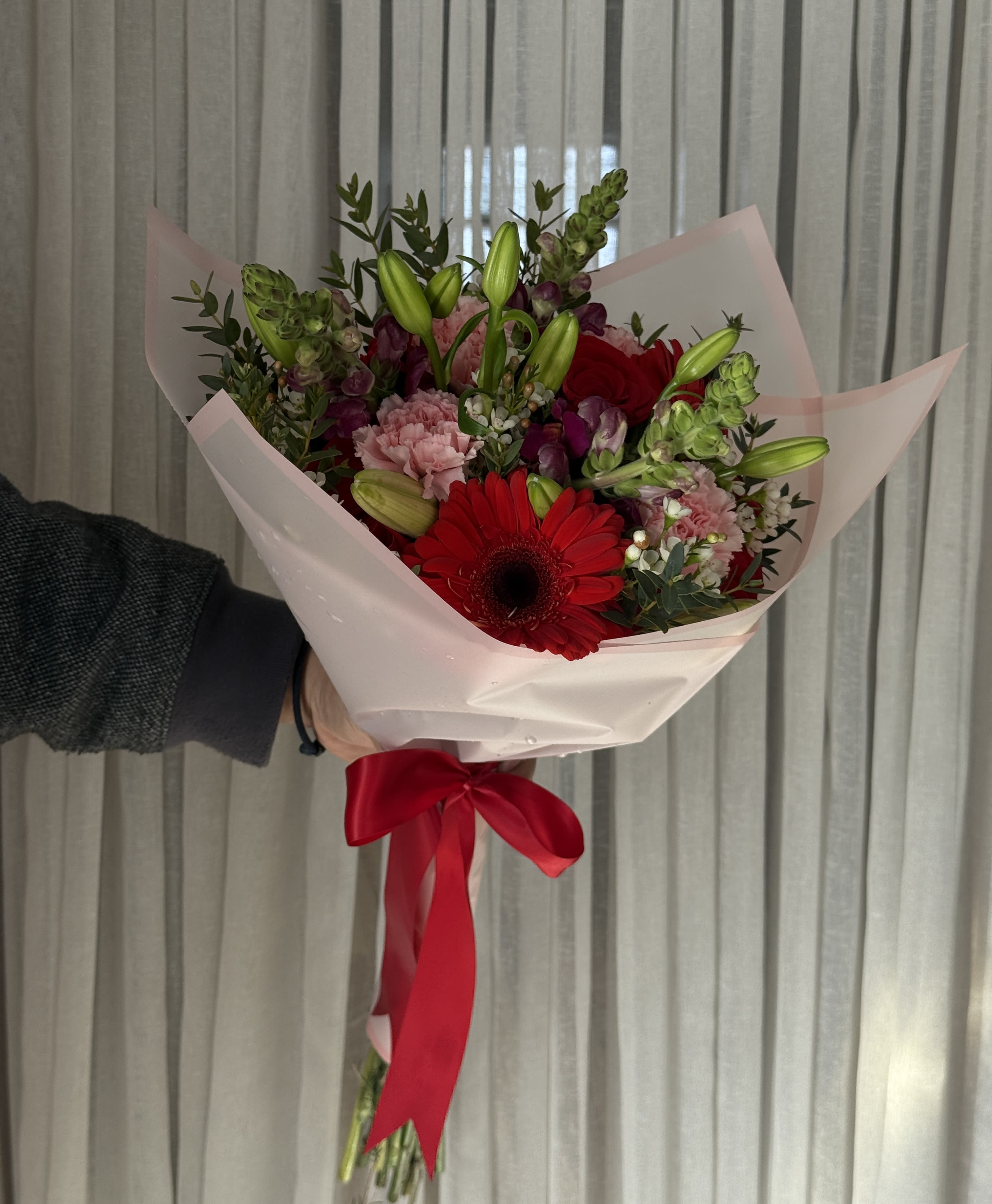 Handheld bouquet of red gerbera daisies, pink carnations, and lily buds wrapped in pale pink paper