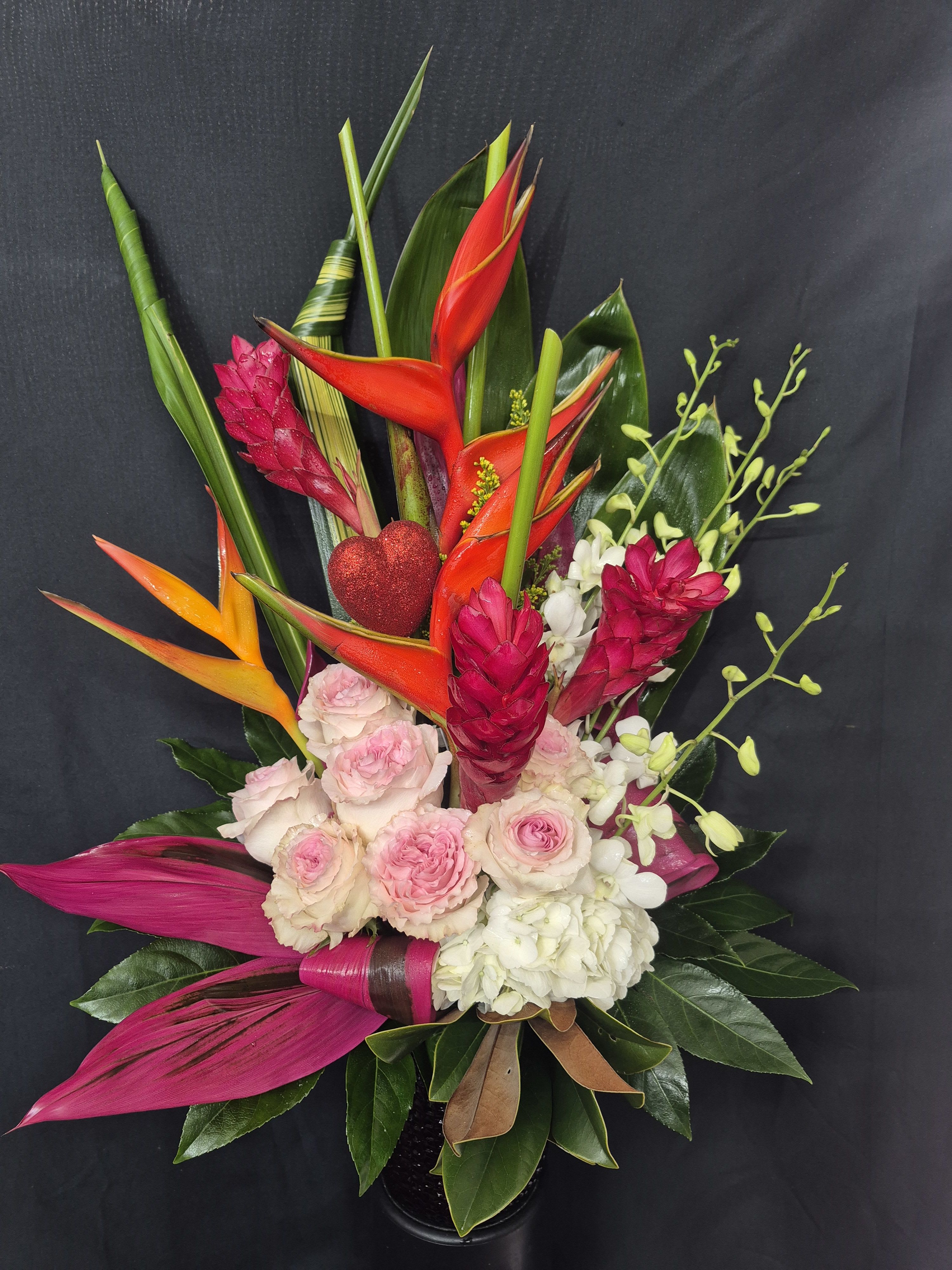 BLUSH AND FIRE - A bold and vibrant tropical arrangement featuring striking heliconia and ginger blooms, complemented by soft blush pink roses and delicate white accents. Designed with lush greenery and a touch of romance,this eye-catching piece blends exotic flair eith elegant charm-perfect for celebrations, love, or making a lating impression.