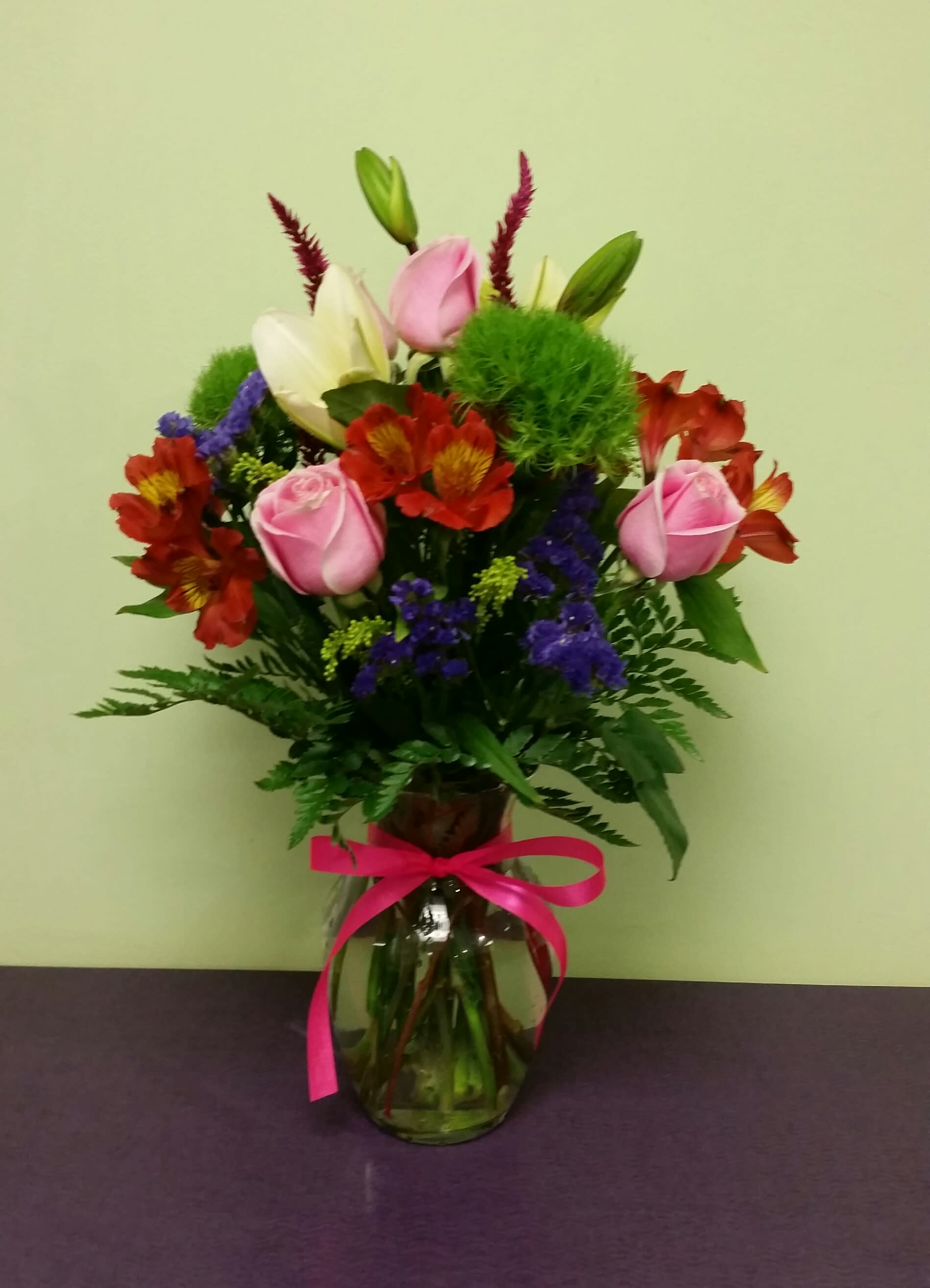 Mixed bouquet of pink roses, lilies, and red flowers in a glass vase with a pink ribbon.