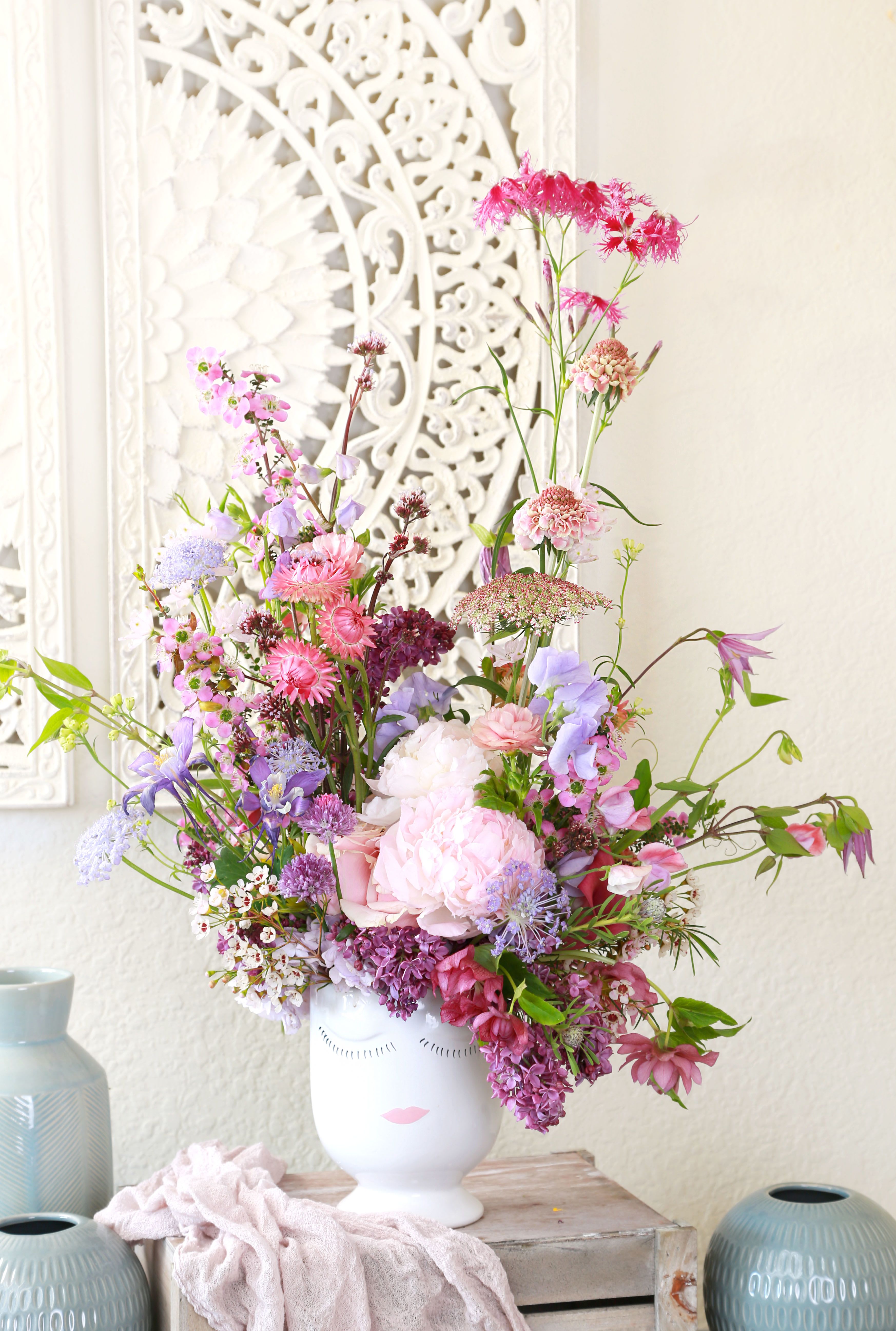 Tall pastel pink and purple flower arrangement in a white ceramic face vase on a rustic crate