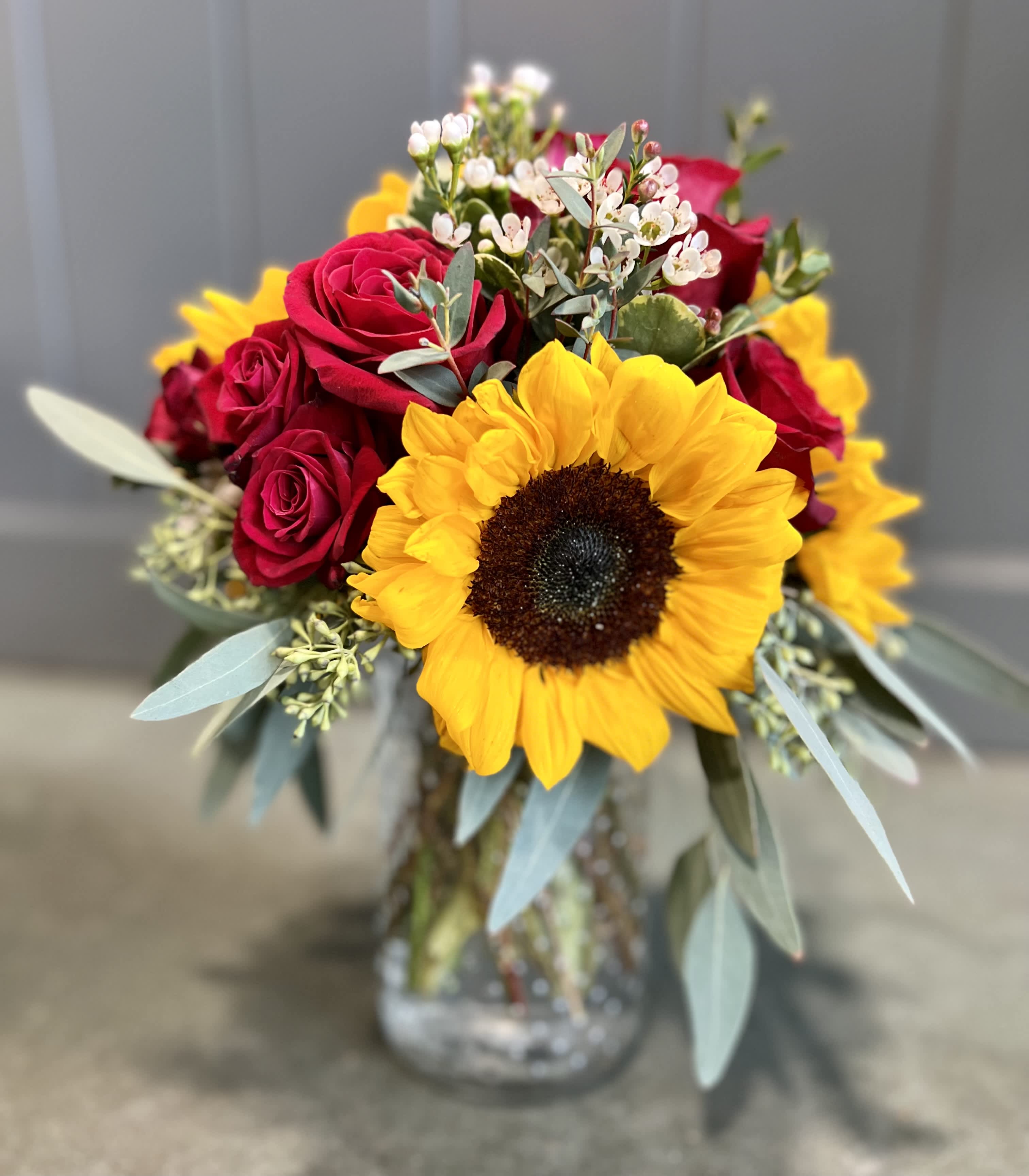 Arrangement of yellow sunflower, red roses, and small white blooms in a clear glass jar vase