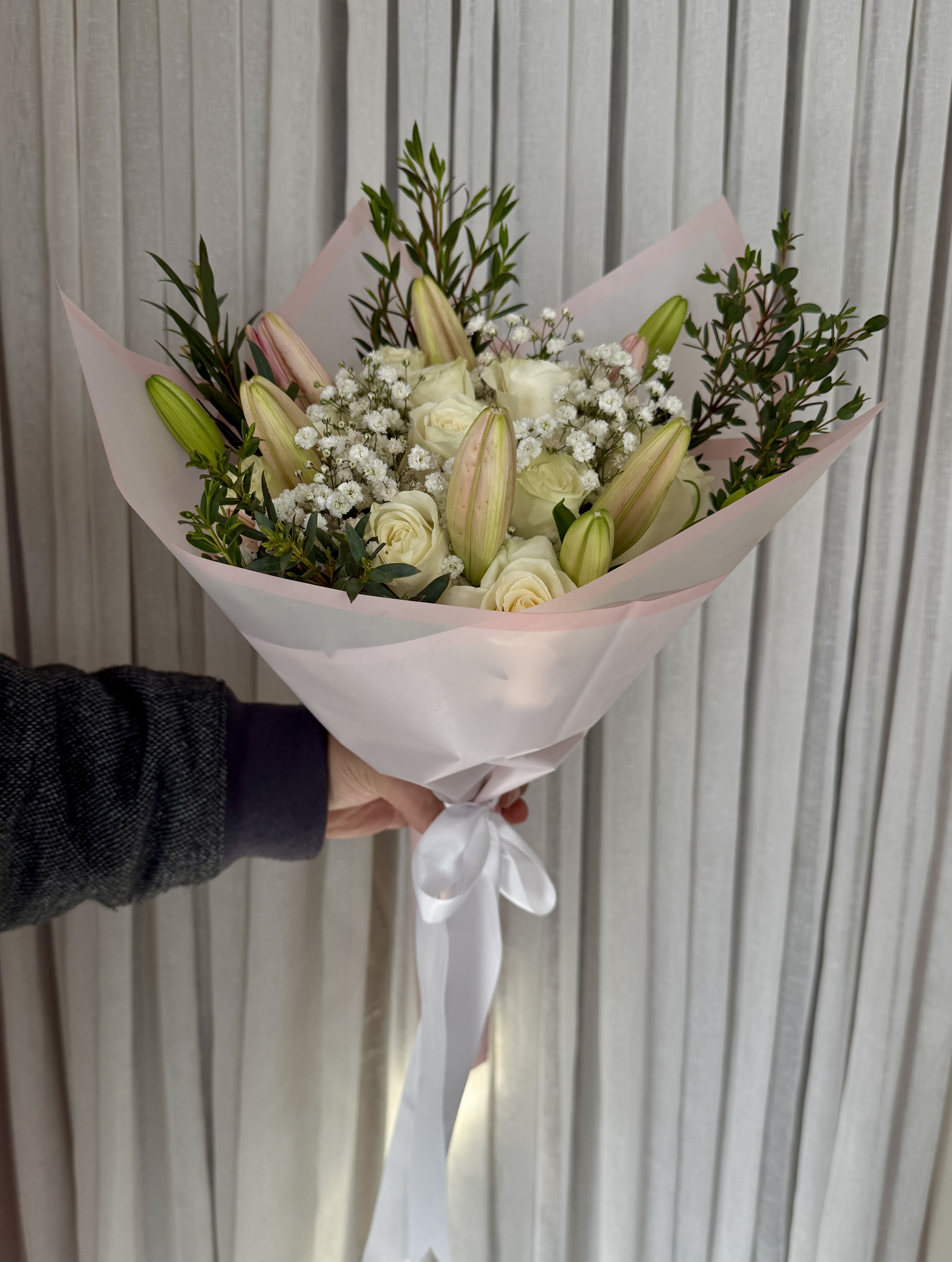Handheld bouquet of white roses and lily buds wrapped in pale pink paper