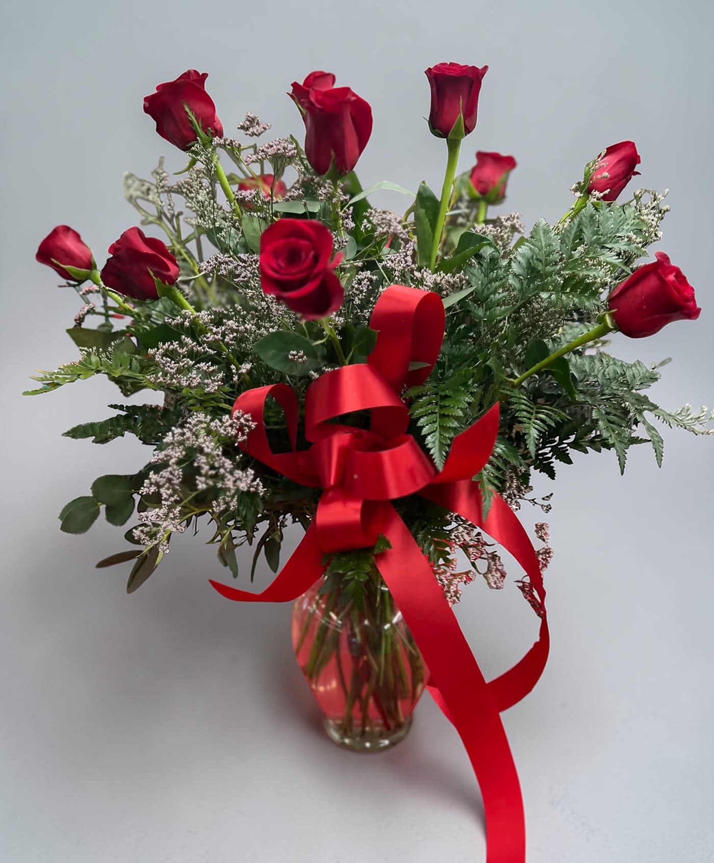 Dozen Roses  - Deep, romantic red roses arranged in a light, hand-tied style make a timeless statement for anniversaries, Valentine's Day, or any occasion when you want someone to feel truly cherished. This dozen-rose arrangement also works beautifully for birthdays, Mother's Day, graduations, "I'm sorry" gestures, or simply to say "thinking of you"-the mix of red, pink, white, and green tones keeps it versatile and season-spanning.  A clear glass vase holds twelve roses set at varying heights with delicate white filler blooms and mixed greenery for an airy, natural silhouette. A large satin red ribbon is tied around the center and drapes down the vase, adding a romantic, celebratory accent that suits weddings, prom, or holiday tables. The presentation is elegant enough for Christmas and New Year gatherings and warm enough for get-well, thank-you, or just-because deliveries.  Designed to make the recipient feel seen and celebrated, this arrangement is a classic choice for love and romance, Mother's Day, Thanksgiving, or welcoming a new baby. Vase-arranged for immediate display, it arrives ready to brighten a room-refresh vase water and trim stems periodically to extend bloom life.