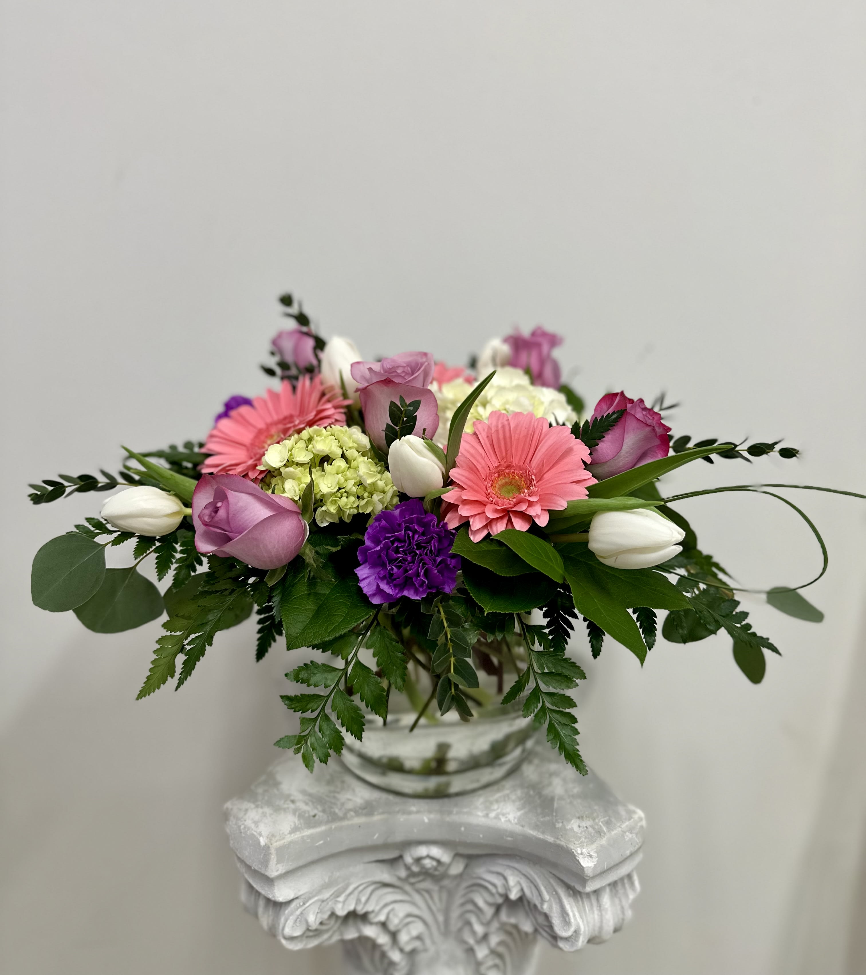Low glass vase arrangement with pink gerbera daisies, purple roses, white tulips, and green hydrangea