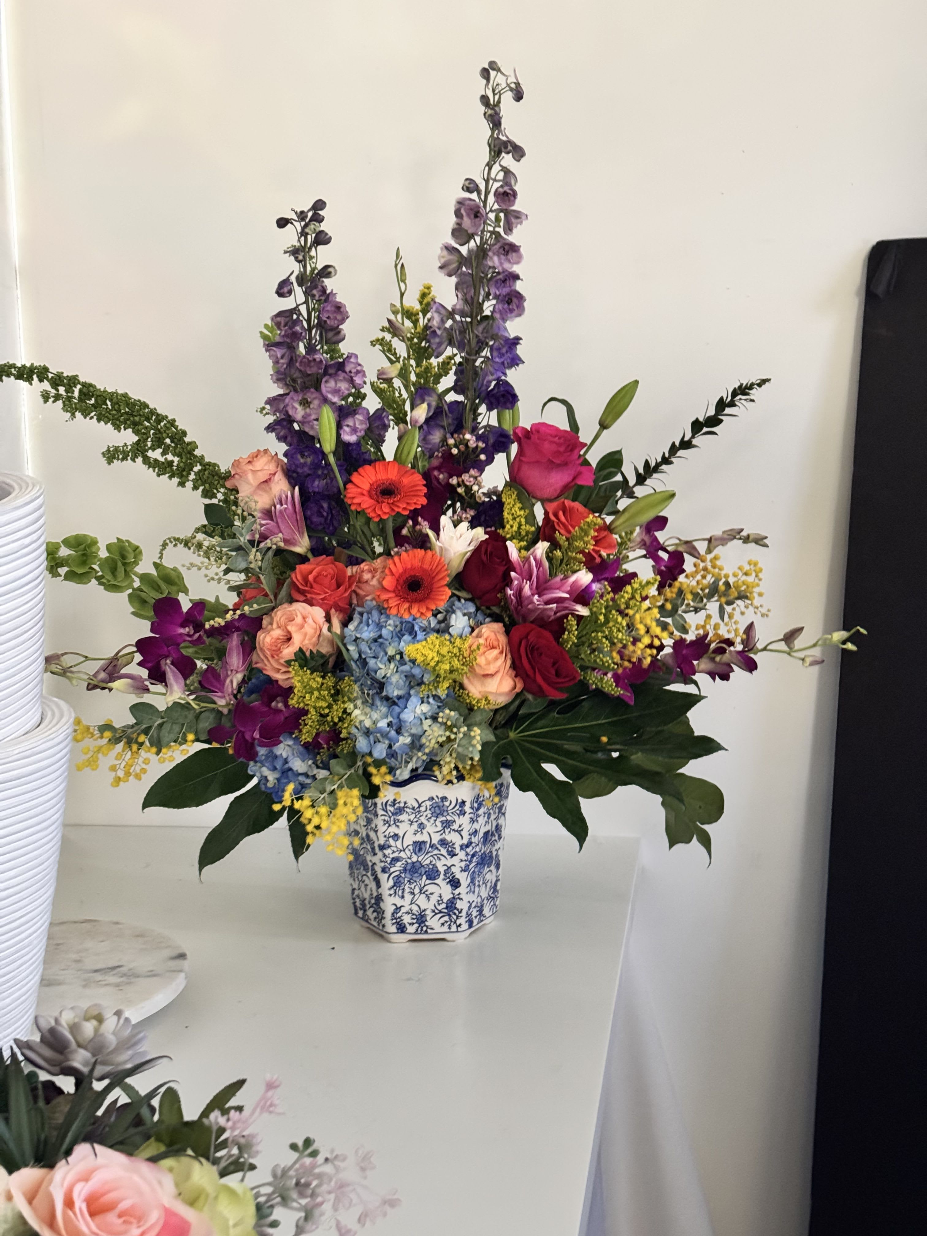 Blooming Bright - A tall, joyful statement in purple, pink, blue and yellow-this flower arrangement is perfect as a birthday bouquet or cheerful celebration gift that makes the person you're thinking of feel bright and loved. The design pairs lush garden textures with airy upward stems so the recipient sees a dramatic, festive silhouette from every angle.  Roses, hydrangeas, gerbera daisies, delphinium, orchids, lilies and alstroemeria are layered into a dense center with lighter stems reaching outward for movement and height. Soft blue hydrangea and vivid purples sit beside hot pinks and coral tones, while bright yellow filler adds lively contrast. The flowers arrive in a blue-and-white ceramic vase that gives classic decorative weight to the vibrant mix.  Ideal for birthdays, congratulations or any moment you want to celebrate, this arrangement reads as both exuberant and thoughtfully composed-she'll feel genuinely celebrated when it arrives. The composition is tall and abundant; vase included for immediate display. For best enjoyment, trim stems and change water every 2-3 days and keep away from direct heat.