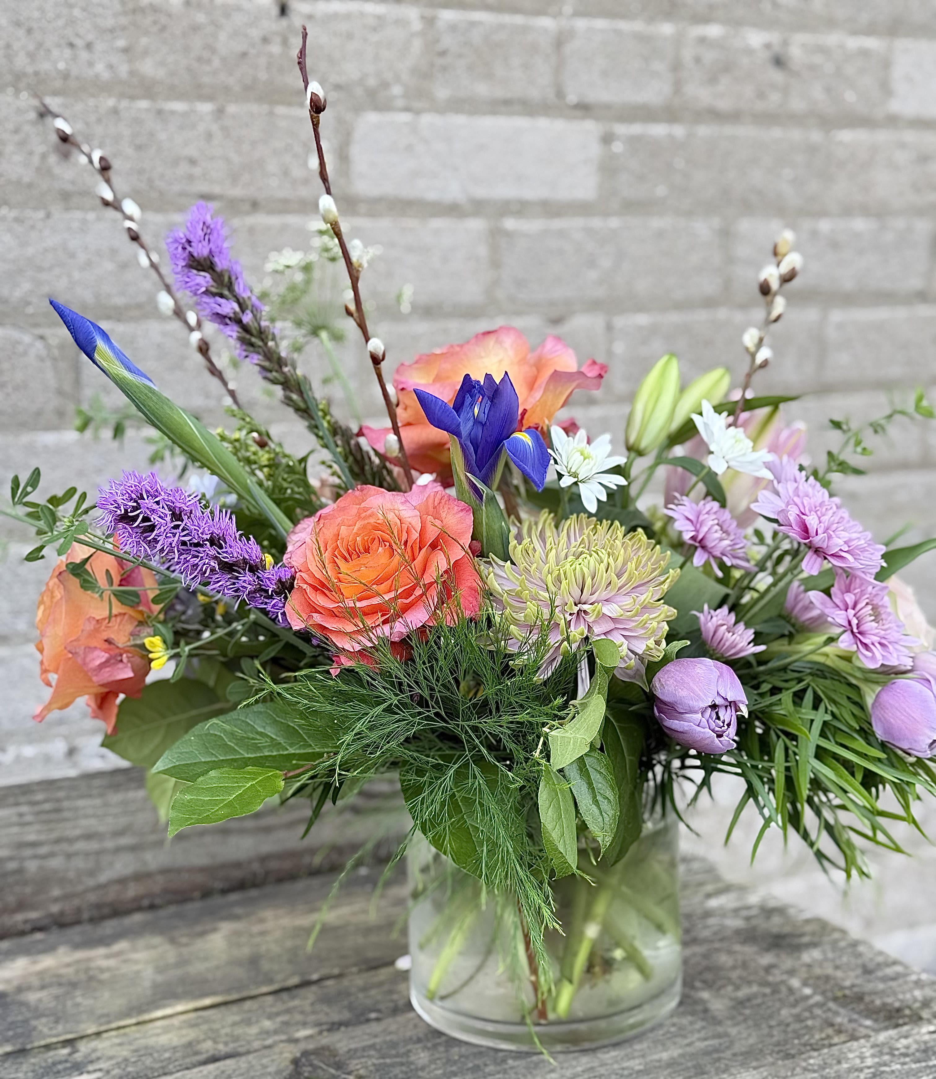 Spring Passion - Coral roses and sky-blue iris mingle with lavender blooms and white daisy-like flowers in an airy, garden-style flower arrangement-an irresistible mixed color spring bouquet perfect for birthdays, thank yous, or a just-because moment. The soft mix of pinks, purples, and blues creates a cheerful, spring-forward impression that will make the person you're thinking of feel truly celebrated and seen.  This loose, natural design features roses, iris, lilies, tulips, chrysanthemums and other seasonal stems arranged with tall, branching lines and varied textures for a lively, casual look. Pale lily buds and daisy-like blossoms add lightness while bright coral and violet tones provide striking contrast. The flowers arrive arranged in a clear glass vase that keeps the presentation simple and fresh; the overall feel is garden-picked and effortless.  Ideal for brightening a kitchen table, office desk, or bedside, this bouquet suits recipients who love soft color palettes and a relaxed, natural style. Standard sizing fits the pictured arrangement-if you need a larger statement or special delivery instructions, please select your preferred size and delivery options at checkout.