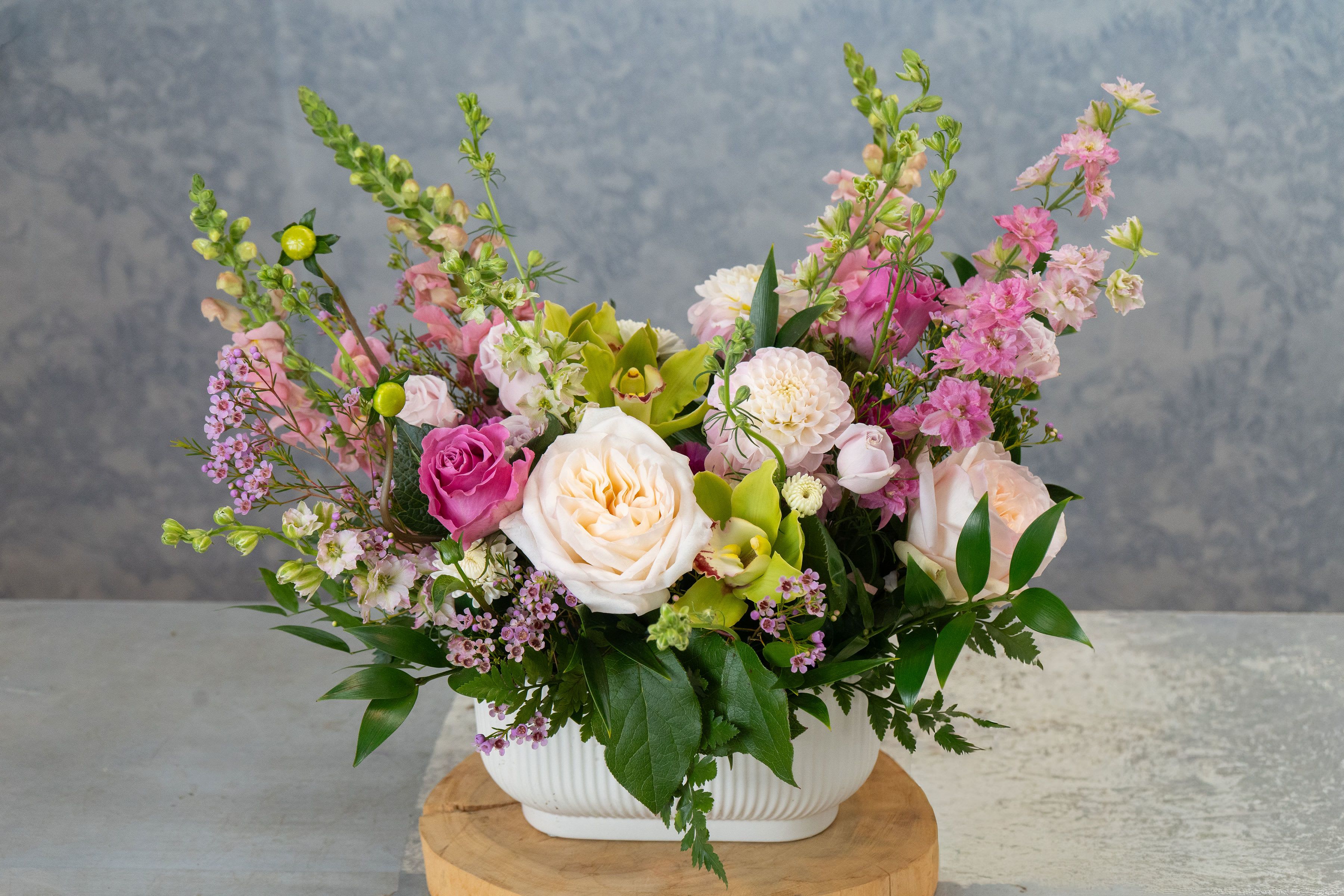 Victoria's Garden - Cream and blush roses mingle with bright green orchids and tall spires of pink snapdragons in this garden-style flower arrangement - for your children's mom, or your mom, or your grandma! Soft white fever few, lavender stock and pom‑pom chrysanthemums lift the composition while glossy foliage and purple accents give the design a modern pop, so the person you're thinking of will feel both cherished and delighted.  Arranged in a simple white ceramic vase, the composition is full and layered: large ivory and blush roses anchor the center, surrounded by dahlias, carnations, spray roses and clustered filler blooms for texture and depth. Tall accent stems rise above the vase for an airy, elegant silhouette while denser lower clusters create a lush, wrapped-in-bloom look. Colors are soft pink, white and lavender with vivid green orchids providing striking contrast.  Perfect for birthdays, anniversaries, spring celebrations or as a meaningful "thinking of you" gift, this arrangement presents as a timeless, celebratory bouquet that suits both intimate and formal settings. Vase‑ready and arranged for lasting impact, it's designed to be enjoyed immediately - trim stems if needed and refresh water daily to keep blooms looking their best.