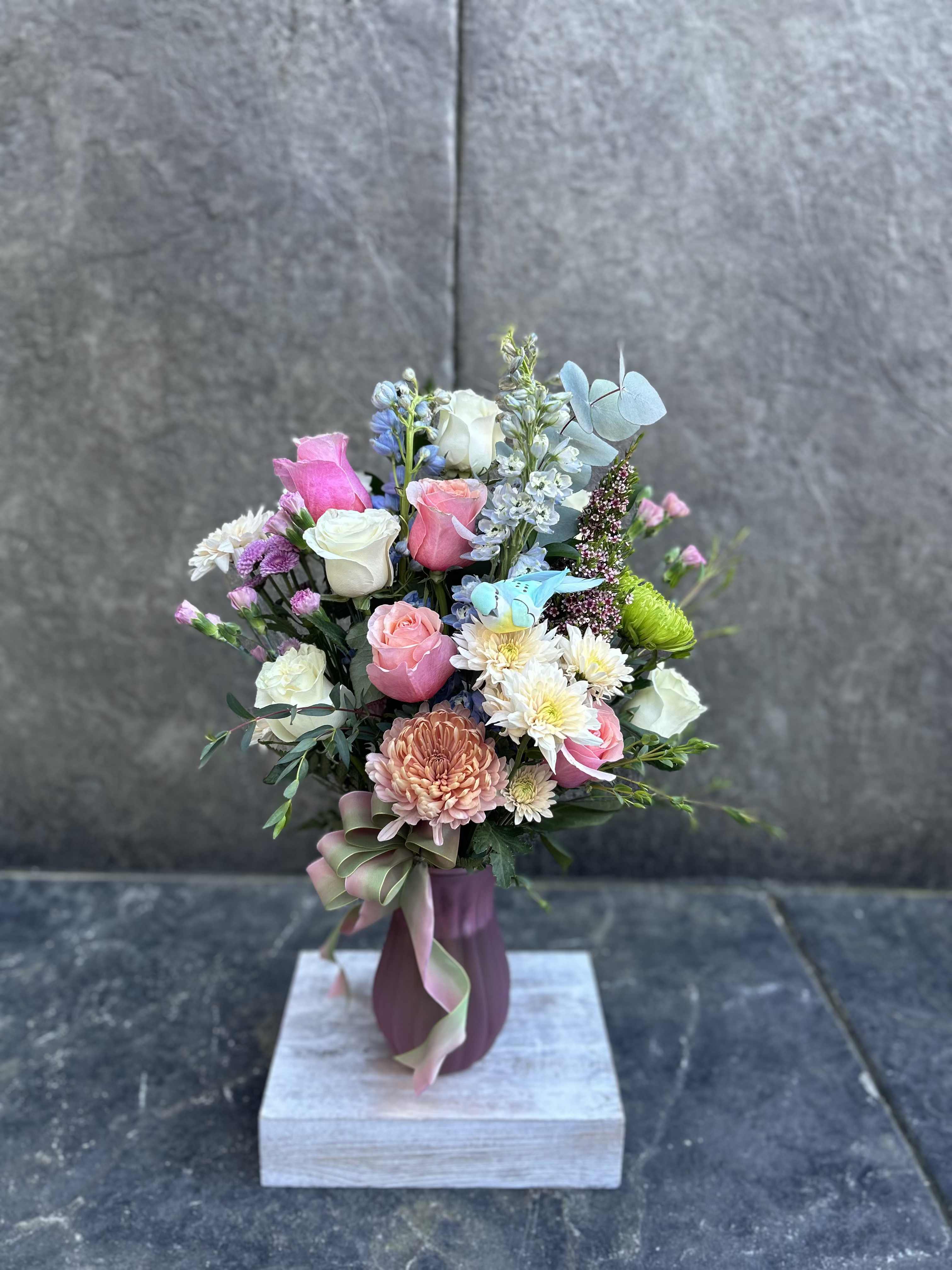 A little bird told me - Pink roses, white roses, delphiniums, chrysanthemums, mini carnations, blooming filler with eucalyptus in a matte pink vase. Decorated with a little pastel bird and ribbon.   *Reasonable substitutions may be made due to floral availability. Will have similar look and feel.