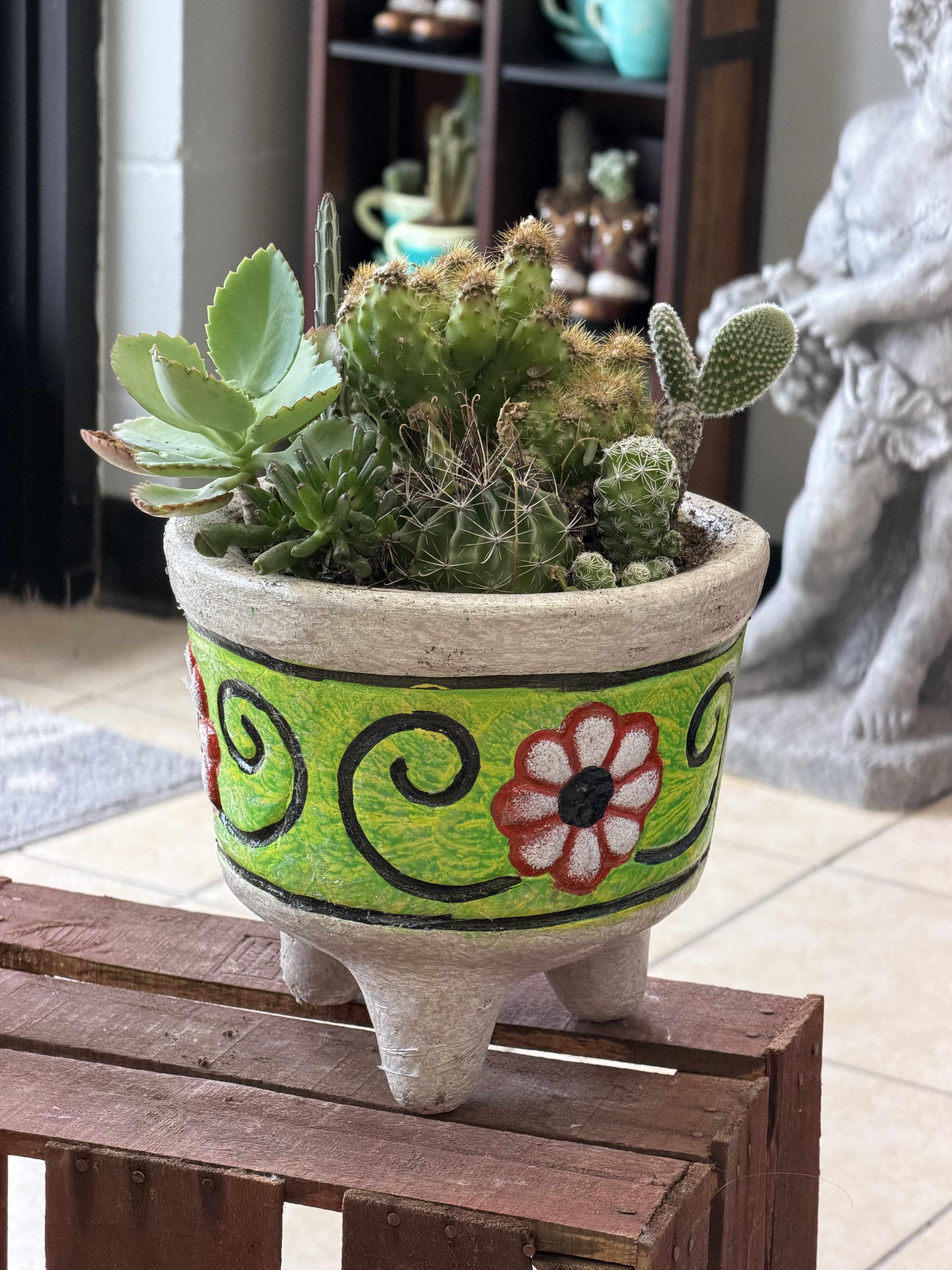 molcajete de suculentas - A hand-painted ceramic planter filled with a sculptural mix of succulents and small cacti makes a lively housewarming or Mother's Day gift that lasts. This succulent arrangement is rooted in a bright green molcajete-style planter decorated with black swirls and red-and-white floral motifs, so it reads as a playful, folk-art statement piece that will catch the eye on a tabletop or windowsill. For birthdays, thank-yous, or a "just because" surprise, the recipient will enjoy a living gift that feels personal and thoughtfully chosen.  The plants themselves range from broad, fleshy-leaved succulents to rounded, spiny cacti, giving a pleasing contrast of textures and forms in shades of green with touches of red, pink, and white from the accent plants and painted details. Planted densely for a full, sculptural look, the arrangement arrives ready to display in its painted ceramic planter-no extra potting required.  Ideal for someone who prefers low-maintenance greenery, this molcajete of succulents brightens a kitchen counter, office desk, or entry table and sends a cheerful, long-lasting message. Water sparingly, place where it receives bright, indirect light, and it will stay healthy with minimal care-perfect for gifting when you want the kind of present that continues to bring a smile.
