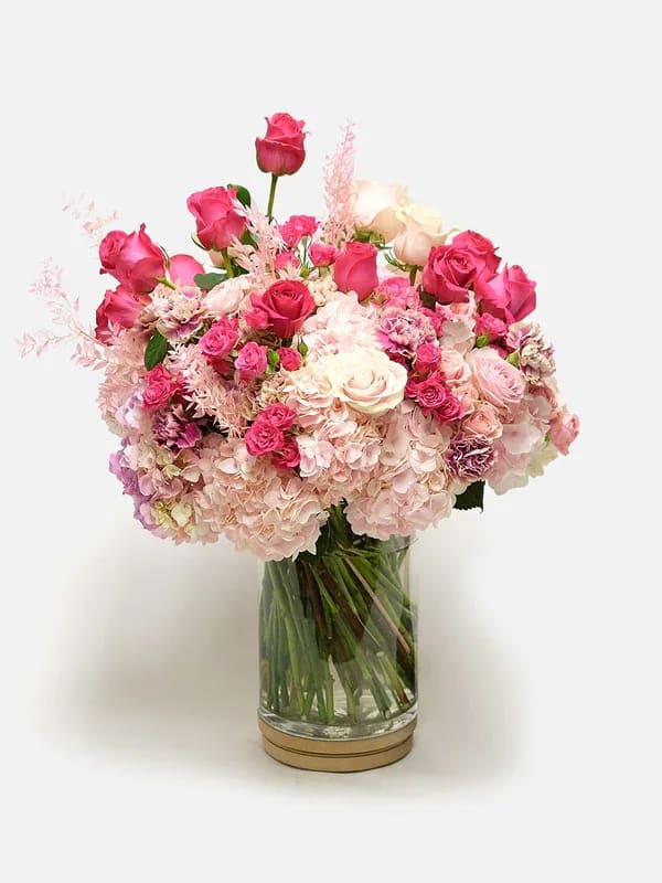 Blush Noir - A lush, feminine arrangement that reads like a love note in bloom - Blush Noir is a romantic flower arrangement perfect for anniversaries, spring celebrations, or any moment you want to say "I love you." Layered pink and white roses create an immediate visual centerpiece while pale pink hydrangeas form a soft, cloud-like base; the overall effect is intimate and celebratory, the kind that makes the person you're thinking of feel truly seen.  This bouquet mixes roses, hydrangeas, and carnations in shades of pink, white, and hints of purple, with small clustered blooms and airy pink filler woven through for texture and movement. Flowers arrive arranged in a clear cylindrical glass vase with a simple round base, offering a polished, classic presentation that suits a dinner table, bedside, or office desk. The dense, compact styling keeps the arrangement looking full and abundant from every angle.  Ideal for expressing love and romance - especially during spring or on an anniversary - Blush Noir sends a message of warmth and devotion. Vase is included; to keep blooms fresh, change the water every other day and recut stems if needed.
