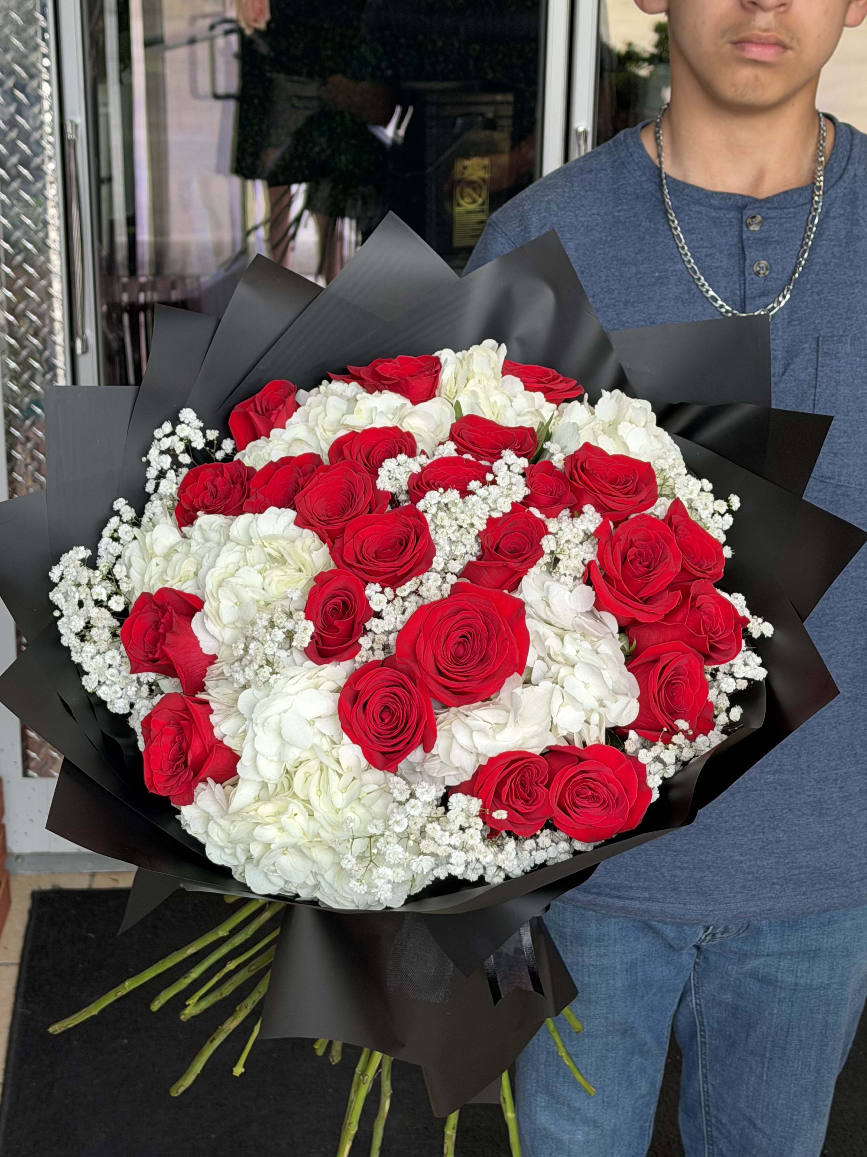 red rose love  - A lush cascade of vivid red roses takes center stage in this red rose love flower arrangement, the perfect anniversary bouquet or Valentine's Day gift to make someone feel deeply adored. Bright white hydrangeas and delicate white filler flowers soften the intensity, while the black wrapping paper provides a dramatic, modern contrast that heightens the romance and polish of the design. This is the kind of bouquet that reads as both luxurious and intentional-ideal for celebrating a milestone, saying "I love you," or marking a special romantic occasion.  Tightly packed and substantial in size, the arrangement features clusters of red roses surrounded by creamy white hydrangea blooms and small white accents for texture and movement. The black wrap frames the flowers like a velvet backdrop, making the reds appear richer and the whites crisper; it also lends a formal, gift-ready presentation that needs no extra adornment. Hand-delivered and arranged upright, this bouquet arrives ready to impress and to be placed in a tall vase for display.  Designed to make the recipient feel truly seen and celebrated, this arrangement suits anniversaries, Valentine's Day, or any moment when you want to declare deep affection. For best enjoyment, refresh the stems in clean water upon arrival and recut stems if you transfer to your own vase; its substantial scale makes it a standout centerpiece as well as a personal gift.