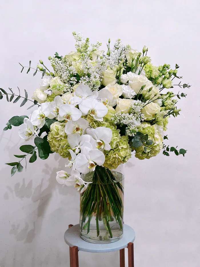 Celeste Bloom - A lush, romantic bouquet of mixed pinks and lavenders that reads like a garden in full bloom-perfect as a birthday bouquet or anniversary roses gift for the person you want to make feel truly cherished. The arrangement centers on soft pink roses and pale blush hydrangeas, with small clustered blooms in pink and lavender adding texture and a few taller rose stems creating graceful height and movement. That feminine, full-rounded silhouette delivers an immediate impression of warmth and celebration.  For a calmer, more refined option-ideal for weddings, sympathy, or a sophisticated congratulations-choose the airy white-and-green composition of white phalaenopsis orchids, pale green hydrangeas, and white roses. This version cascades with long stems and eucalyptus-like foliage for an elegant, loose shape that reads both fresh and formal. Both designs arrive in a clear glass vase (the white-and-green style shown displayed on a round stool), making them easy to place and enjoy; choose the colorway that best suits the moment and the person you're honoring.