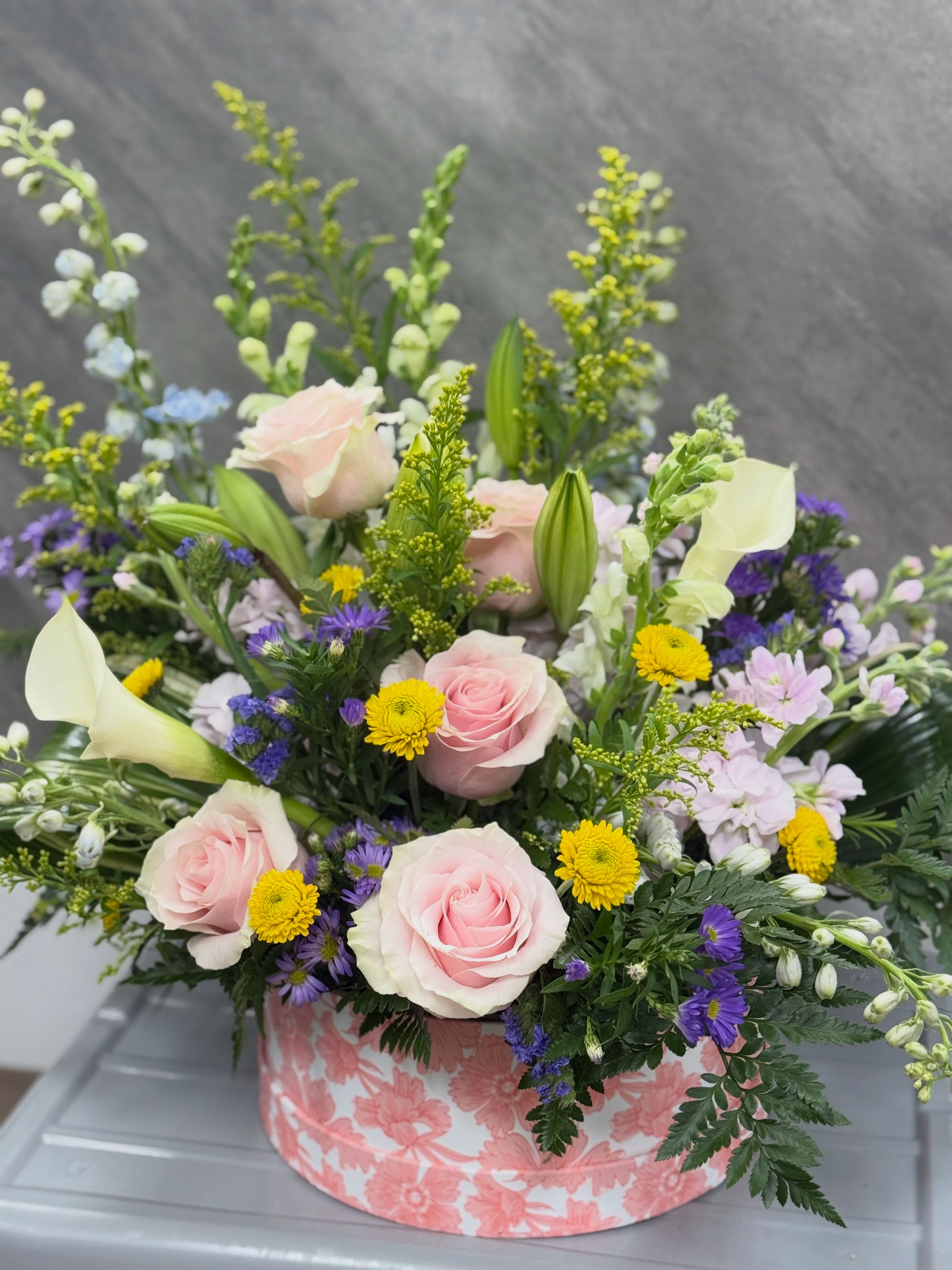 Soft pink - Soft pink roses and white calla lilies create a gentle, garden-fresh look that makes this flower arrangement perfect for a birthday bouquet, a thank-you gift, or any spring celebration. The soft pink, purple, yellow, and white palette reads as cheerful and feminine-an arrangement that will make the person you're thinking of feel seen and celebrated.  Layered and full in a low, rounded floral box with a pastel pink floral print, the design mixes roses, calla lilies, chrysanthemums (yellow button mums), asters and snapdragons for texture and movement. Taller accent stems rise behind the main cluster to give a natural, garden-style silhouette while the compact shape keeps it easy to display on a table or desk.  Send this as a thoughtful everyday arrangement to brighten a friend's day, celebrate a mom, or add spring warmth to a hostess gift. The low box makes arranging and hydration simple-refresh the water daily and trim stems if you want to extend vase life-available in one standard size ready for local delivery.