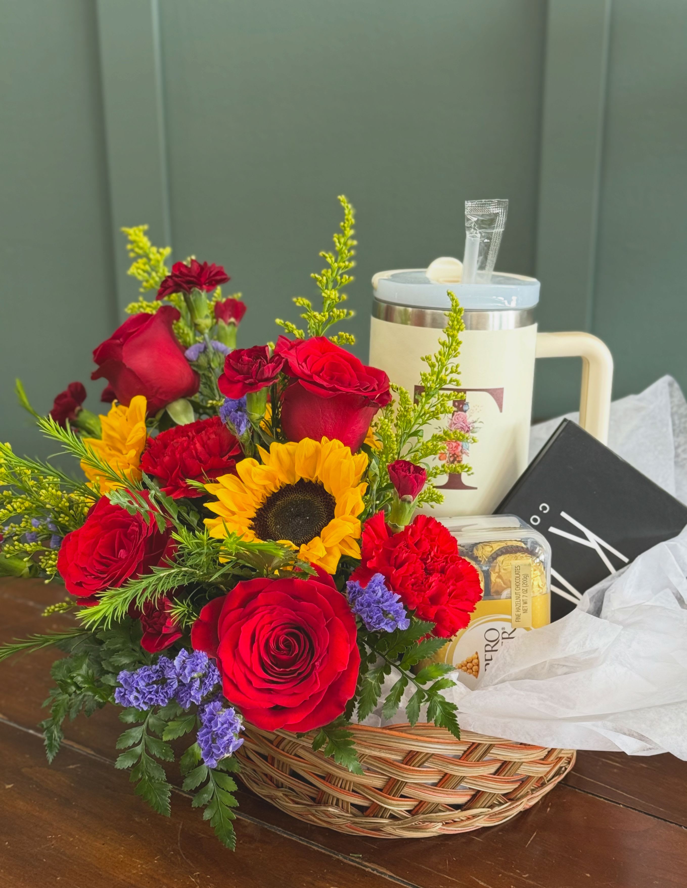 Gift  Basket - Vivid red roses and bright yellow sunflowers take center stage in this gift basket, making it an unforgettable birthday bouquet or a warm thank-you gift. The combination of romantic red and sunny yellow-accented by white and purple blooms-creates a cheerful, celebratory look that also suits anniversaries or a simple "thinking of you" surprise for someone special.  Arranged low in a woven basket, the flowers include red roses, red carnations, sunflowers and small purple filler blooms, layered on tissue paper and wrapped in clear plastic for presentation and transport. A ceramic mug and a box of chocolates are tucked into the arrangement, turning the floral display into a ready-to-give gift set that feels thoughtful and complete.  Perfect for the person who appreciates both beauty and a little indulgence, this basket arrives ready to present-no extra wrapping needed. Size and freshness are maintained by the low-profile design and careful packaging; order now to ensure delivery for birthdays, anniversaries, or to brighten someone's day.