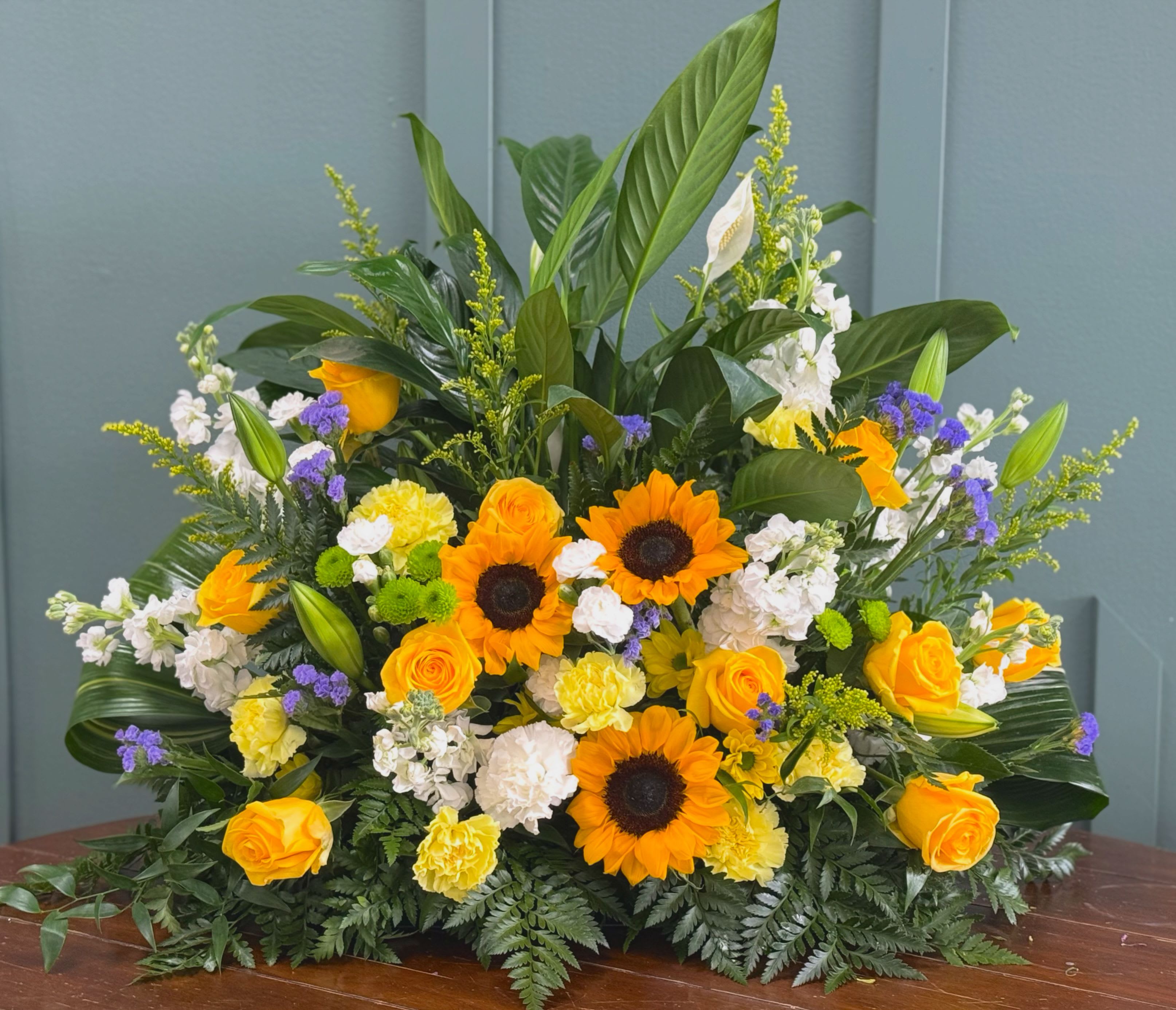 Peace lily  arrangement  - A lush, low centerpiece that brings warmth and calm, perfect as a funeral sympathy arrangement or a bright table focal point for a celebration. This flower arrangement features the contrast of sunny sunflowers and yellow roses with serene white stock and pale carnations, so the person you're thinking of will feel comforted and uplifted at the same time.  The design is full and layered: glossy tall foliage rises behind the blooms while fern-like greenery fans across the base, creating a garden-fresh texture. Pops of small purple accents add contrast to the yellow and orange palette, giving the piece visual depth and a lively, cheerful note without overpowering the calming whites.  Sized as a large, low centerpiece, it sits well on dining or memorial tables and is arranged for immediate display-no vase fussing required if provided in a container. Light daily water refreshes will keep the stock and carnations looking their best; please contact us for delivery timing to ensure it arrives fresh for the occasion.