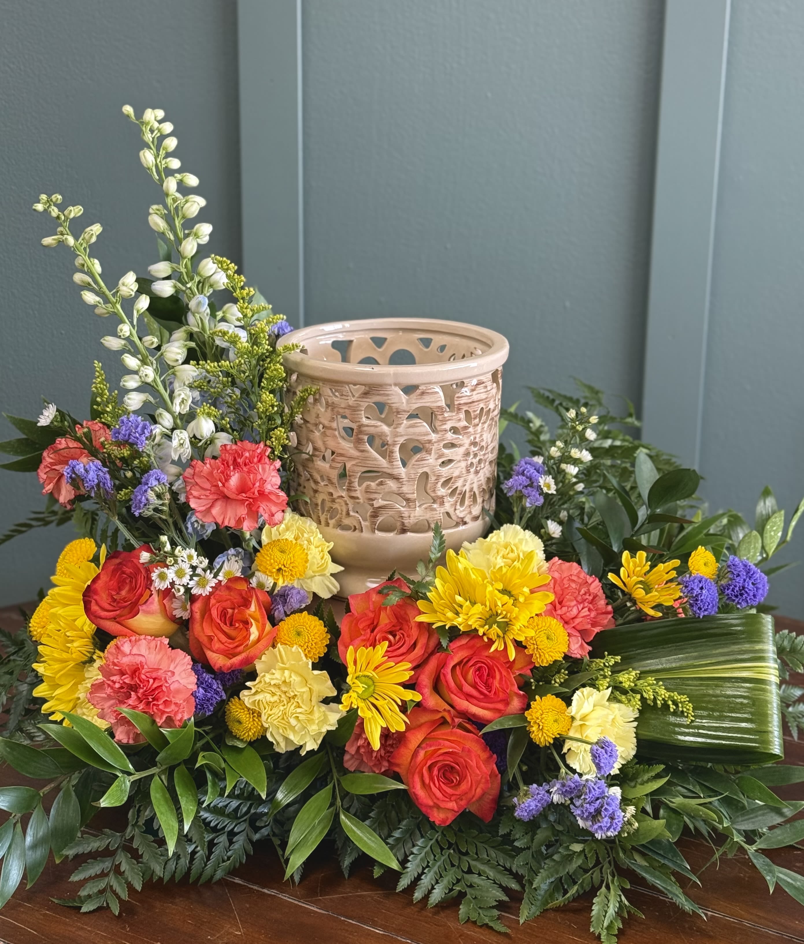 Cremation - A bright, celebratory centerpiece arranged around a decorative candle holder, this cremation flower arrangement is designed as a polished tribute or memorial display that also works beautifully for remembrance ceremonies. The compact, low silhouette puts the candle at the heart of the design so the person you're honoring will be remembered with warmth and color rather than somber tones alone.  Roses, carnations, chrysanthemums and cheerful daisy-like blooms are clustered in vivid pinks, reds, yellows and purples, while tall white snapdragon spikes add vertical rhythm and gentle contrast. The base is finished with mixed greenery and a broad leaf wrap for a lush, formal presentation that reads well from every angle. The arrangement's dense, layered composition gives it a centerpiece quality that feels both respectful and uplifting.  Choose this piece when you want to offer a meaningful, colorful expression of sympathy or to mark a celebration of life - the kind of tribute that helps family and friends remember joyful moments. Its low, finished form is ready to place on a table or altar; keep away from drafts and follow standard candle-safety practices when the decorative candle is lit.