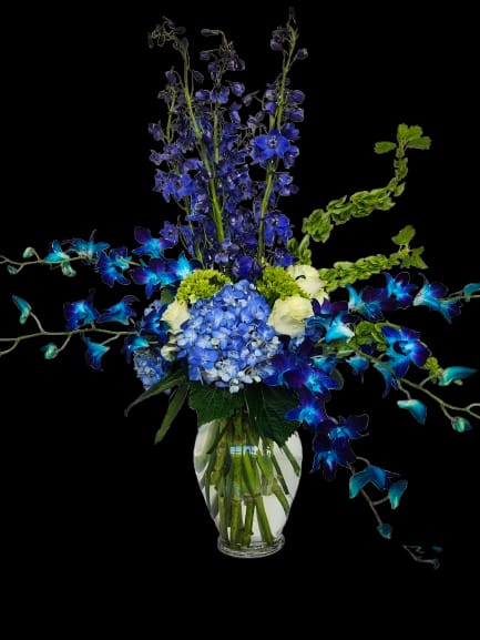 Avatar - A bold blue hydrangea cluster anchors this contemporary flower arrangement, designed to make the recipient feel celebrated. As an everyday bouquet for birthdays, congratulations, or a thoughtful thank-you, the mix of blue, white, green, and purple reads cool and elegant-perfect for someone who loves modern, saturated color and airy silhouettes.  Tall, deep-blue delphinium spikes create vertical drama while bright blue‑purple dendrobium orchid sprays fan outward for a striking, wide profile. White accents and fresh greenery soften the composition, all arranged in a clear glass vase that showcases stems and keeps the focus on the saturated blooms. Sized to sit on a table or desk, this arrangement delivers a refined, eye-catching statement with minimal fuss.
