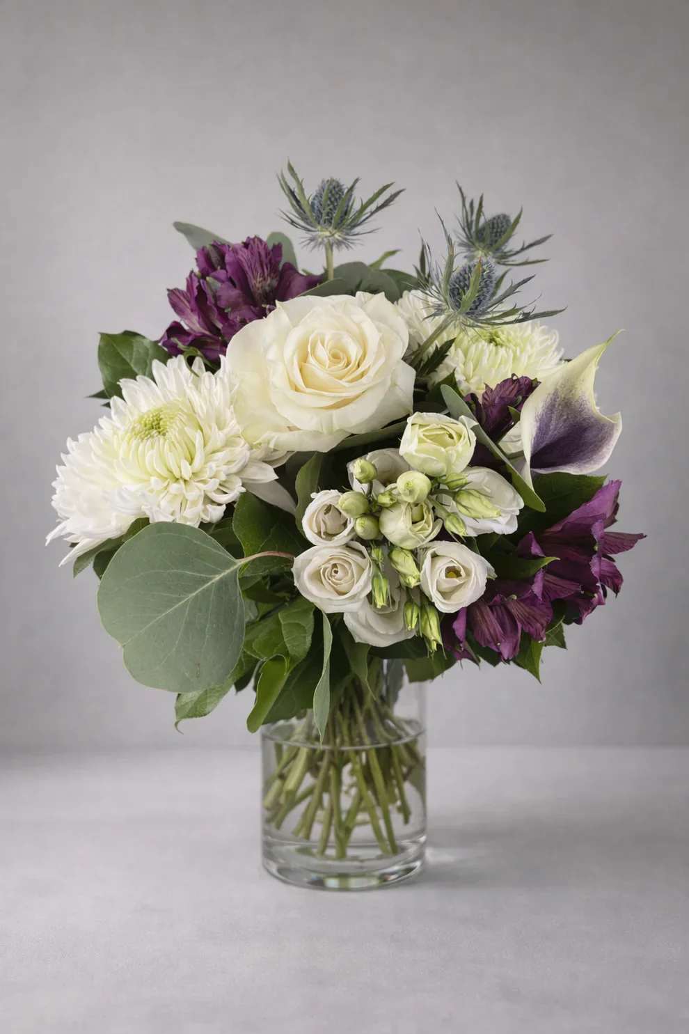 Ethereal Silence - A large ivory rose anchored among white chrysanthemums and a purple-and-white calla lily gives this floral arrangement a quietly dramatic opening - Ethereal Silence is a refined flower arrangement perfect for sympathy, a formal gift, or a low-key celebration. The cool palette of white, purple, and green reads both elegant and comforting, the kind of bouquet that makes the person you're thinking of feel seen and supported without overwhelming the room.  The design pairs soft textures and sculpted lines: a focal ivory rose and clusters of white chrysanthemums sit beside purple Alstroemeria and a striking purple-and-white calla lily, all arranged with broad eucalyptus leaves and other lush greenery in a clear glass vase. Spiky blue thistle-like accents rise above the main cluster to add height and a touch of unexpected texture, creating a balanced silhouette that's as tasteful on a mantel as it is on a memorial table.  Made to convey calm and care, Ethereal Silence suits moments when you want to offer sympathy, show appreciation at a formal occasion, or celebrate quietly. The arrangement arrives in a clear glass vase ready to display; trim stems and refresh water every few days to keep blooms looking their best.
