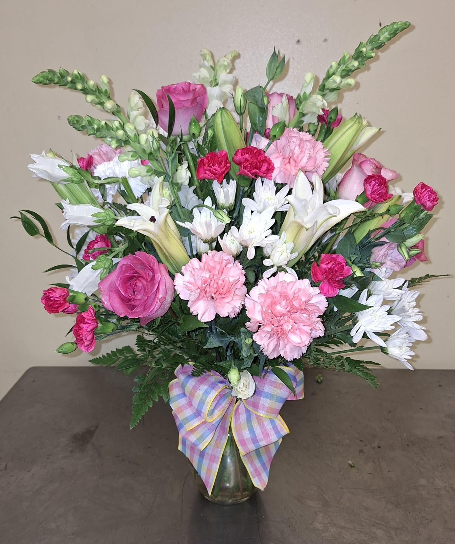 sympathy arrangement - A lush, rounded sympathy arrangement in soft pinks, whites and gentle greens offers a comforting, tender message for someone grieving. This sympathy arrangement places roses, carnations and lilies front-and-center so the recipient feels embraced by classic, fragrant blooms, while the taller snapdragons add a hopeful upward line-appropriate for funeral services, memorial visits, or sending condolences to someone at home.  The clear glass vase holds a full cluster of pastel roses and carnations framed by white lilies and small daisy-like blooms, creating layered texture and soothing contrast. Taller snapdragon spikes rise above the main mass for graceful height, and the pastel plaid ribbon bow at the base gives a soft, handcrafted touch. Colors read as pink, white and green with mixed accents that keep the design gentle yet uplifting.  Thoughtful for sending to a family home, funeral chapel, or as a bedside tribute, this arrangement reads as both respectful and quietly cheerful so the person you're thinking of feels seen and comforted. The glass vase makes it easy to display; water fresh stems as needed and trim stems slightly every few days to prolong bloom life.