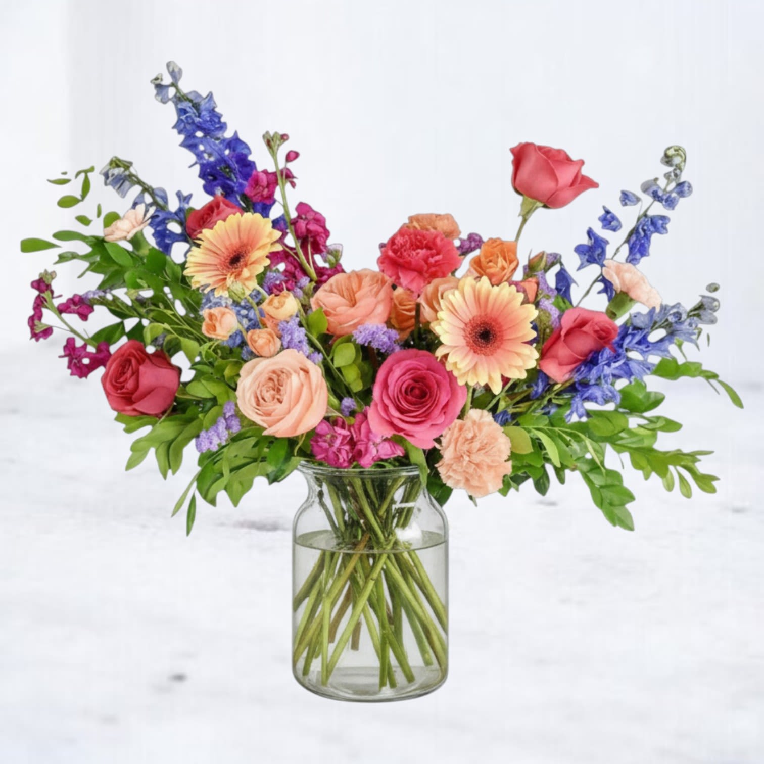 Color Surge - A lively sweep of warm pinks and corals punctuated by cool blue spikes - this garden-style flower arrangement brings an instant upbeat mood to a birthday bouquet or a cheerful "just because" gift. The loose, airy silhouette spreads outward for a casual, romantic feel so the person you're celebrating will feel genuinely seen and brightened by the mix of colors. Peach carnations, coral and pink garden roses, pale gerbera daisies and tall blue delphinium are arranged with soft greenery in a clear glass vase, creating contrast between the warm blooms and cool accents. The composition reads fresh and textural, with airy height from the spiked blooms and a broad, lively shape that looks beautiful on a dining table, desk, or entry console.  Perfect for birthdays, thank-yous, or everyday celebrations, this arrangement arrives ready to display in the included clear glass vase. For best longevity, keep water topped up and trim stems every few days to help the recipient enjoy the colorful display longer.  Whiskey Wallflower is a design-forward florist based in Magnolia, offering same-day flower delivery throughout Magnolia, Montgomery, The Woodlands, Conroe, Tomball, Willis, Spring, and surrounding North Houston communities. Each arrangement is thoughtfully composed using premium blooms, with a focus on modern design, quality, and intentional detail.