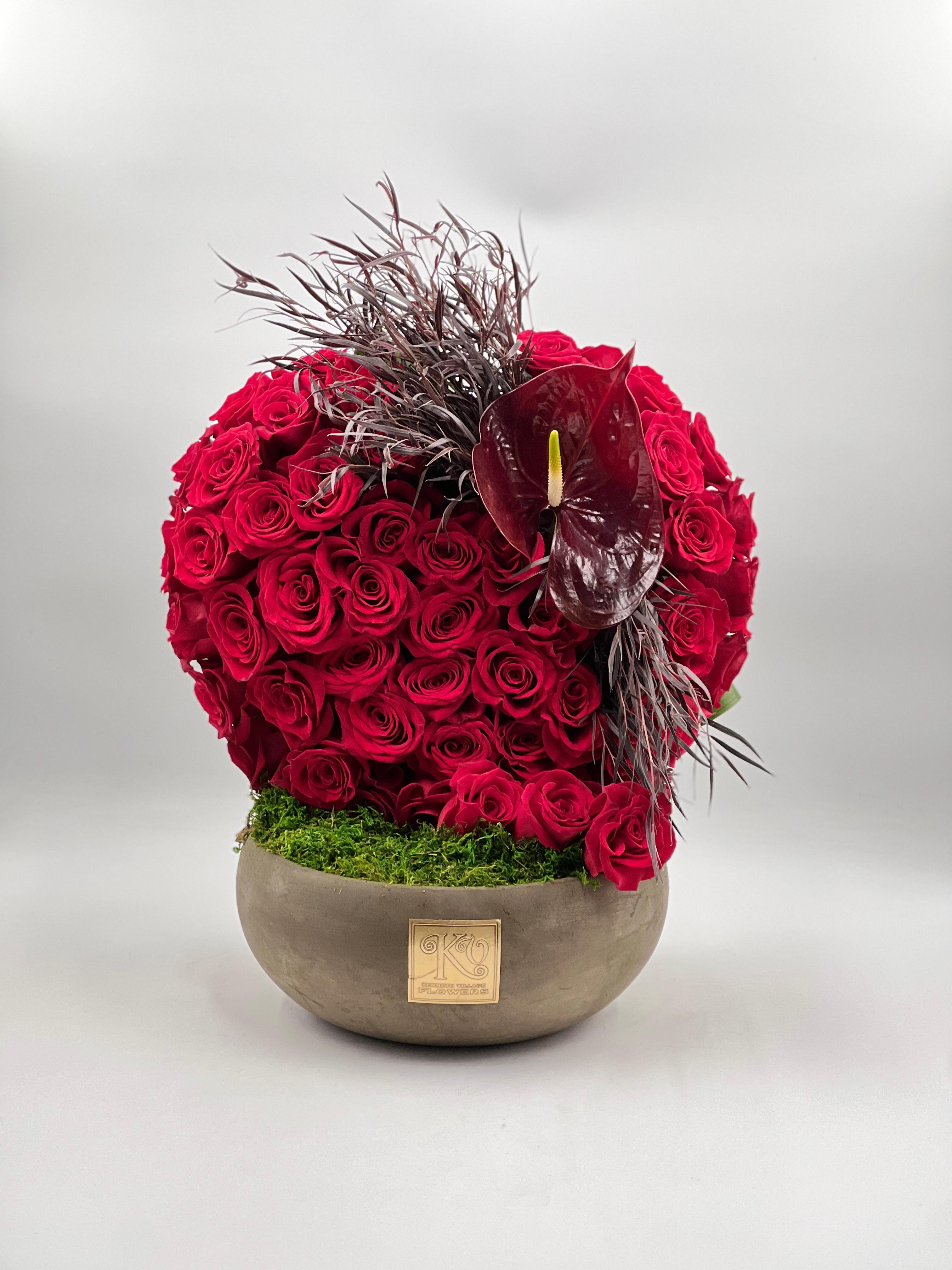 Round arrangement of red roses in a low bowl with a dark anthurium accent