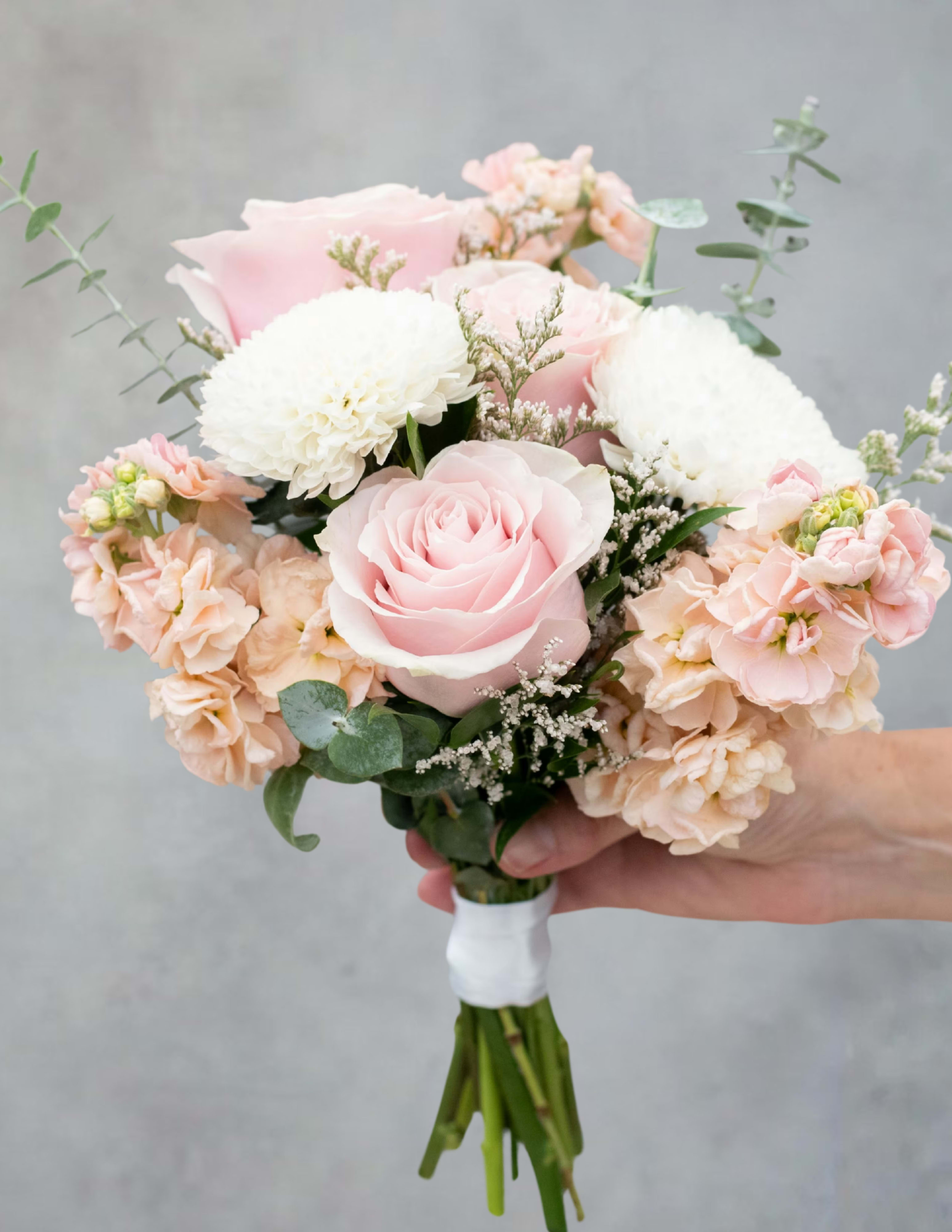 Pastel prom bouquet - Pale pink roses take center stage in this pastel prom bouquet, a soft and romantic choice for prom, weddings, anniversaries, or any sentimental event. The arrangement reads like a garden-picked posy-designed to make the person you're honoring feel cherished and graceful on a special night.  Creamy white chrysanthemums and soft peach carnations surround the roses for a layered, textured look, while small filler blooms and sprigs of eucalyptus add airy movement and muted green contrast. Hand-tied for a natural silhouette, the compact-to-medium bouquet is finished with a neat white wrap at the stems for comfortable carrying and a clean presentation.  Perfect for a corsage alternative, bridal party hand bouquet, or a keepsake for someone who prefers gentle tones, this bouquet is meant to be worn or carried and admired up close. Its fresh, delicate construction keeps blooms looking their best for the event; order with enough lead time so we can deliver the freshest stems for your occasion.