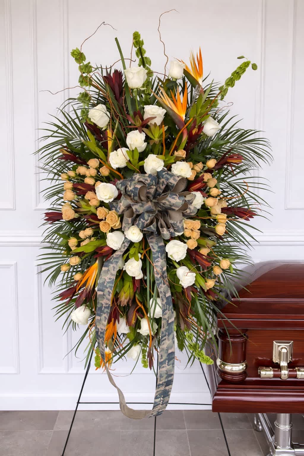 Honor &amp; Valor Tribute - A bold and meaningful tribute, Honor &amp; Valor Tribute is designed to celebrate a life of strength, service, and dedication. This striking standing spray features an artful blend of white roses, peach accents, and exotic tropical elements including birds of paradise, creating a powerful and distinguished presentation.  Lush greenery and dynamic textures give the piece depth and movement, while the camouflage-inspired ribbon adds a deeply personal and symbolic touch—perfect for honoring a veteran, service member, or someone who lived with courage and pride.  Displayed on a standing easel, this arrangement makes a strong and respectful statement at any service or memorial, offering a heartfelt expression of remembrance, gratitude, and honor. For order customization please call the flower shop.