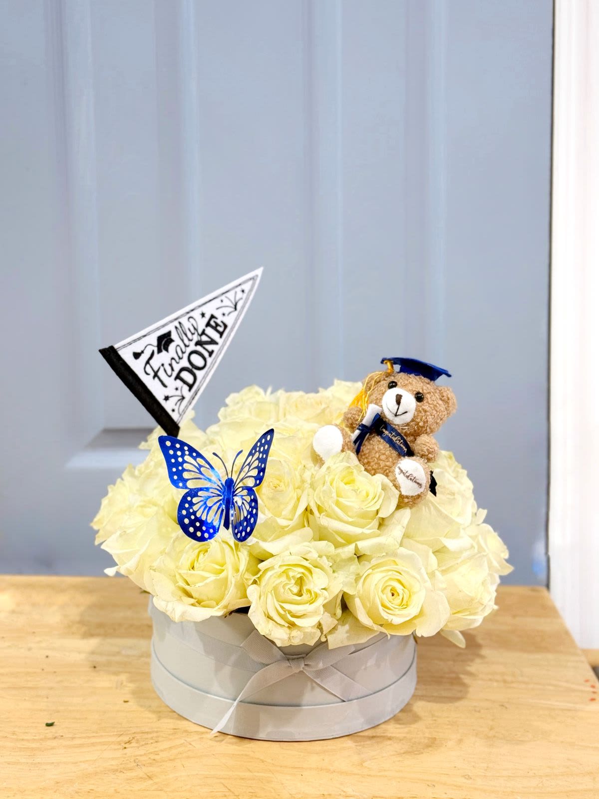 Cream rose arrangement in a round box with a teddy bear and graduation flag