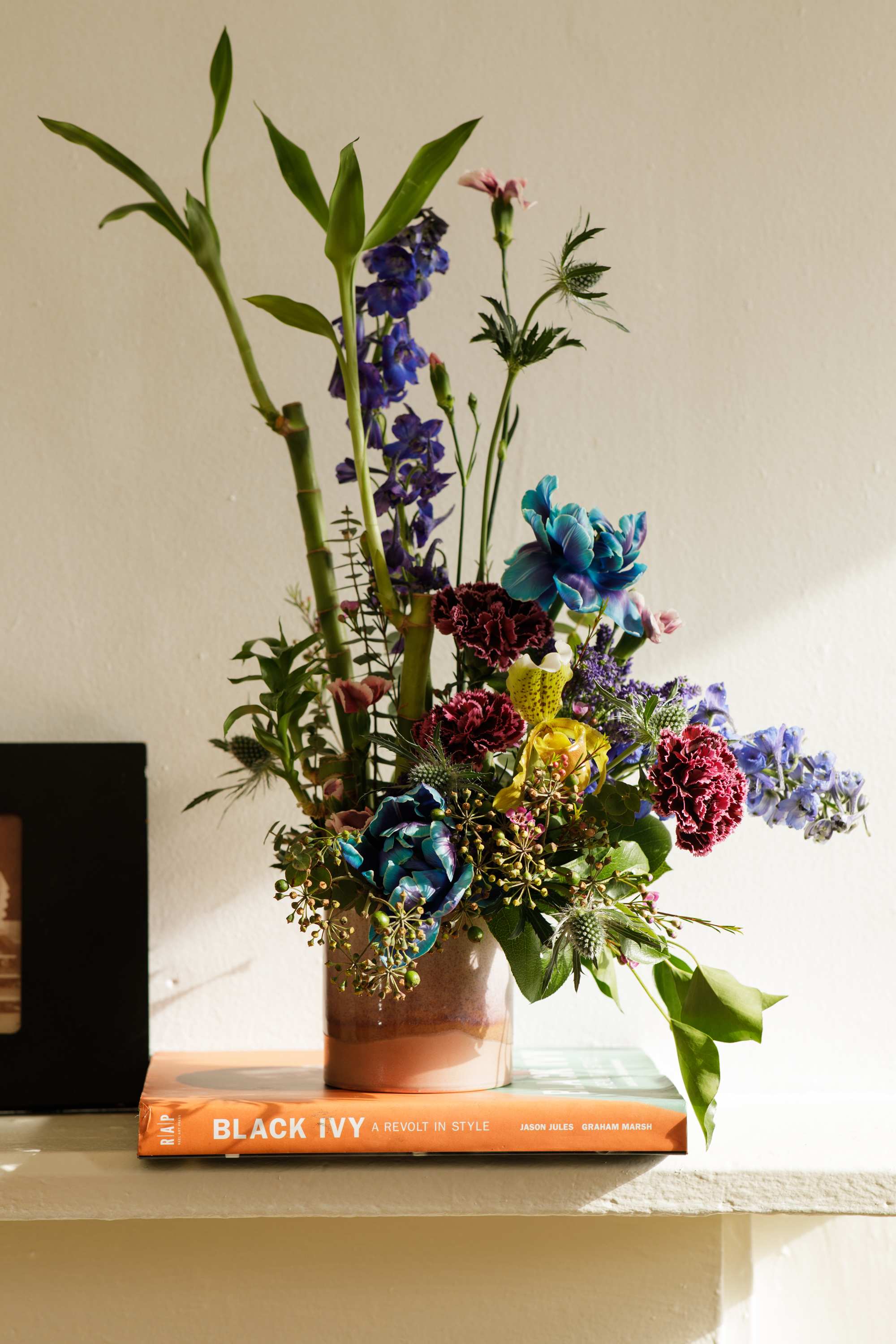 Tall mixed flower arrangement with blue, red, and yellow blooms in a ceramic vase on an orange book.
