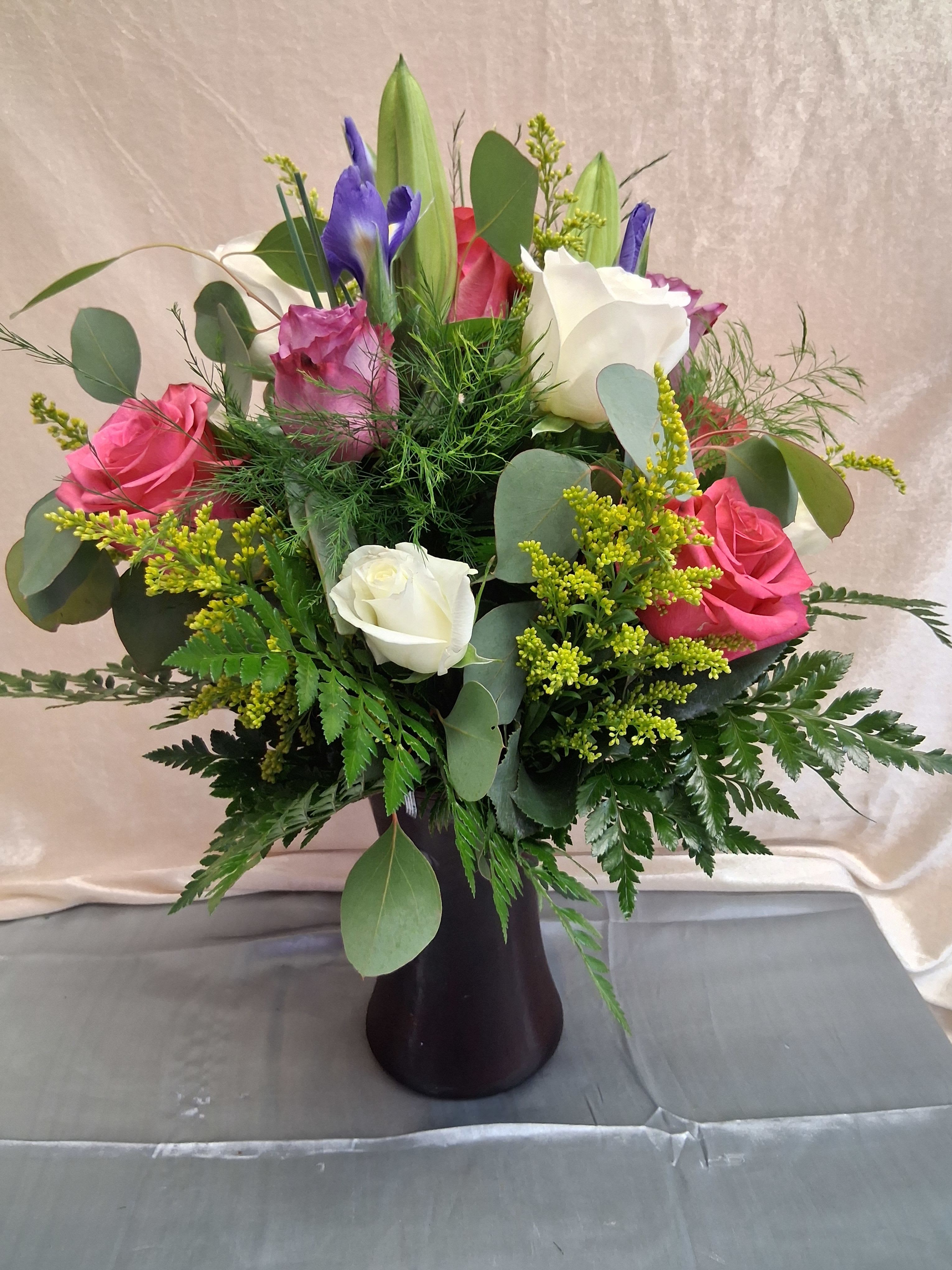 Elegant arrangement with mixed colored roses and greenery - Soft pink and creamy white roses mingle with striking purple irises and tall, unopened lily buds to create a romantic anniversary bouquet that also works beautifully for birthdays or when you want to let someone know you're thinking of them. The upright, layered composition gives the arrangement a graceful, slightly formal presence-she'll feel truly celebrated by the mix of tender rose petals and the sculptural iris forms.  This flower arrangement includes roses, irises, lilies, dense greenery, and airy filler to add volume and texture. Colors run from white and pink to purple and fresh green, arranged in a balanced, vertical silhouette. The stems are presented in a simple dark vase that grounds the design and complements the soft blooms for a refined, gift-ready display.  Perfect for marking anniversaries, milestone birthdays, or offering sympathy with elegance, this bouquet suits recipients who appreciate classic romance with a modern edge. Stems arrive arranged in the vase for immediate enjoyment; trim stems and refresh water every few days to extend vase life and keep the lilies and roses looking their best.