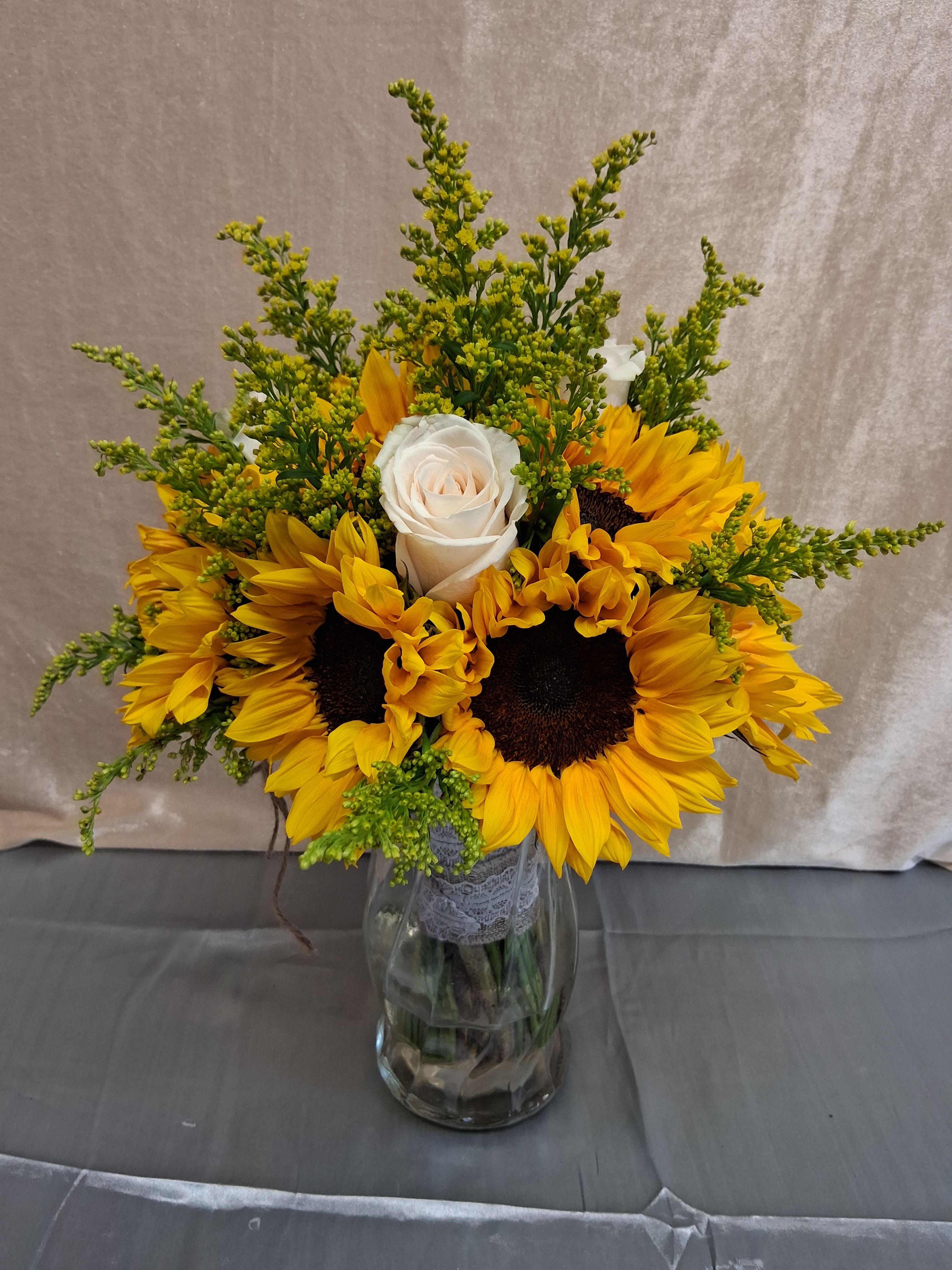 Sunflower Wedding Bouquet  - Bright, sunny sunflowers clustered around white roses to create a cheerful wedding bouquet that also works beautifully for birthdays, thank-you gifts, or a just-because surprise. The contrast of bold yellow petals and the quiet white rose at the center gives the recipient an immediate feeling of warmth and celebration-perfect for a bride who loves rustic charm or for someone you want to make feel genuinely seen on their special day.  This flower arrangement pairs large, vibrant sunflowers with a solitary white rose and wispy green filler sprays for a full, airy silhouette. Stems sit in a clear glass vase wrapped with delicate lace ribbon, blending country elegance with a light, joyful palette of yellow, white, and green. The mix of textures-the sunflower's coarse centers, the rose's soft petals, and the fine greenery-creates visual depth while keeping care simple.  Ideal for a casual or outdoor wedding, a cheerful anniversary, or brightening someone's week, this bouquet sends a message of warmth and appreciation. Vase-ready and hand-tied with lace, it arrives arranged and ready to display; refresh the water every few days and trim stems for the longest-lasting blooms.