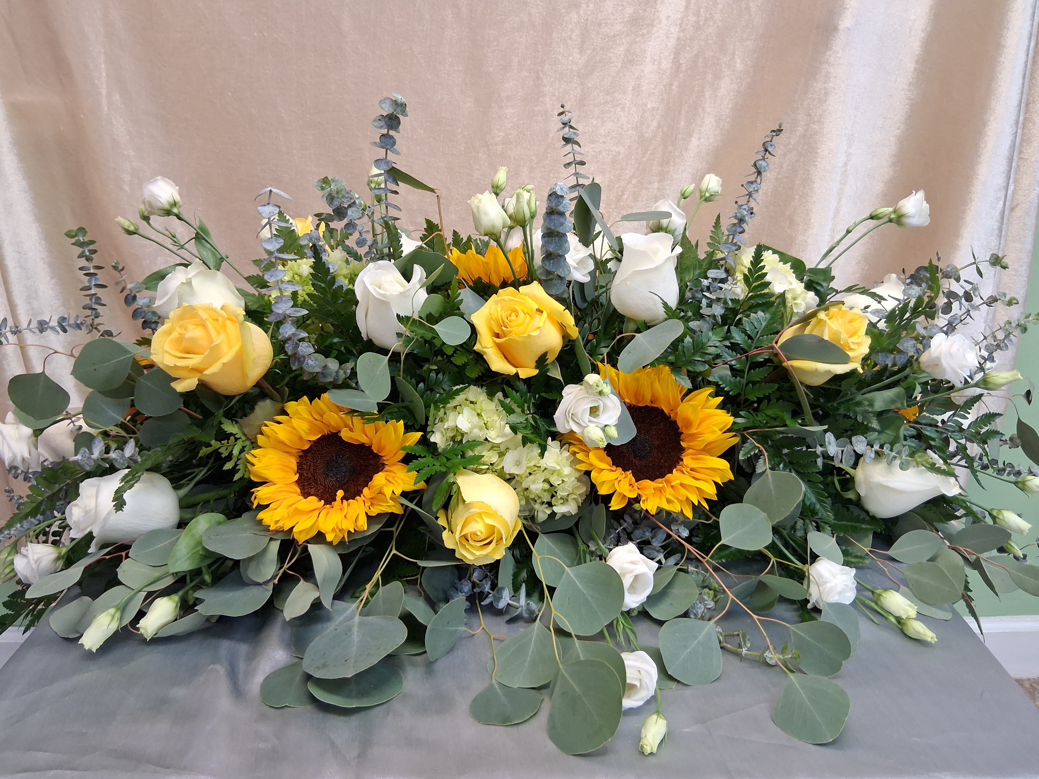 Elegant sunflower center piece for all occasions  - A wide, low centerpiece bursting with sunflowers and yellow roses sets a warm, celebratory tone-perfect as a birthday bouquet, wedding reception table arrangement, or a thoughtful sympathy display. The arrangement's garden-style sweep reads as generous and welcoming, so the person you're honoring will feel seen whether this marks a joyful gathering or a gentle remembrance.  This mixed-color design pairs bright sunflowers and yellow roses with white lisianthus, hydrangea accents, calla lilies, and other seasonal blooms, all framed by blue-gray eucalyptus and layered greenery for an airy, organic silhouette. The composition is large and low, sprawling across the table rather than standing tall, and comes finished with a coordinating table covering to anchor the display.  Sized to make an impact on dining or event tables, this centerpiece delivers a fresh, cheerful color story and a soft texture from its foliage. It's the kind of floral gift that fills a room with warmth-order early for events to ensure availability and the most vibrant, in-season blooms.