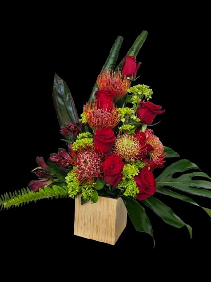 Protea - A dramatic floral arrangement centered on spiky orange protea-like blooms and deep red roses that create a bold, sculptural statement - perfect as a birthday bouquet or romantic anniversary arrangement. The strong color contrast of red, orange, pink accents and bright green filler flowers reads vivid and energetic, so the person you're surprising will feel celebrated and seen the moment the box is opened.  Large tropical leaves fan outward to give the design height and a lush, sculptural silhouette, while a modern wooden cube vase grounds the composition with natural texture. The mix of roses, protea-like flowers, and greenery makes this a standout gift for milestone celebrations, congratulations, or a heartfelt "thinking of you" moment; the sturdy vase also makes it easy to display on a dining table or office desk. For best longevity, keep in fresh water and out of direct heat to enjoy the arrangement for days.