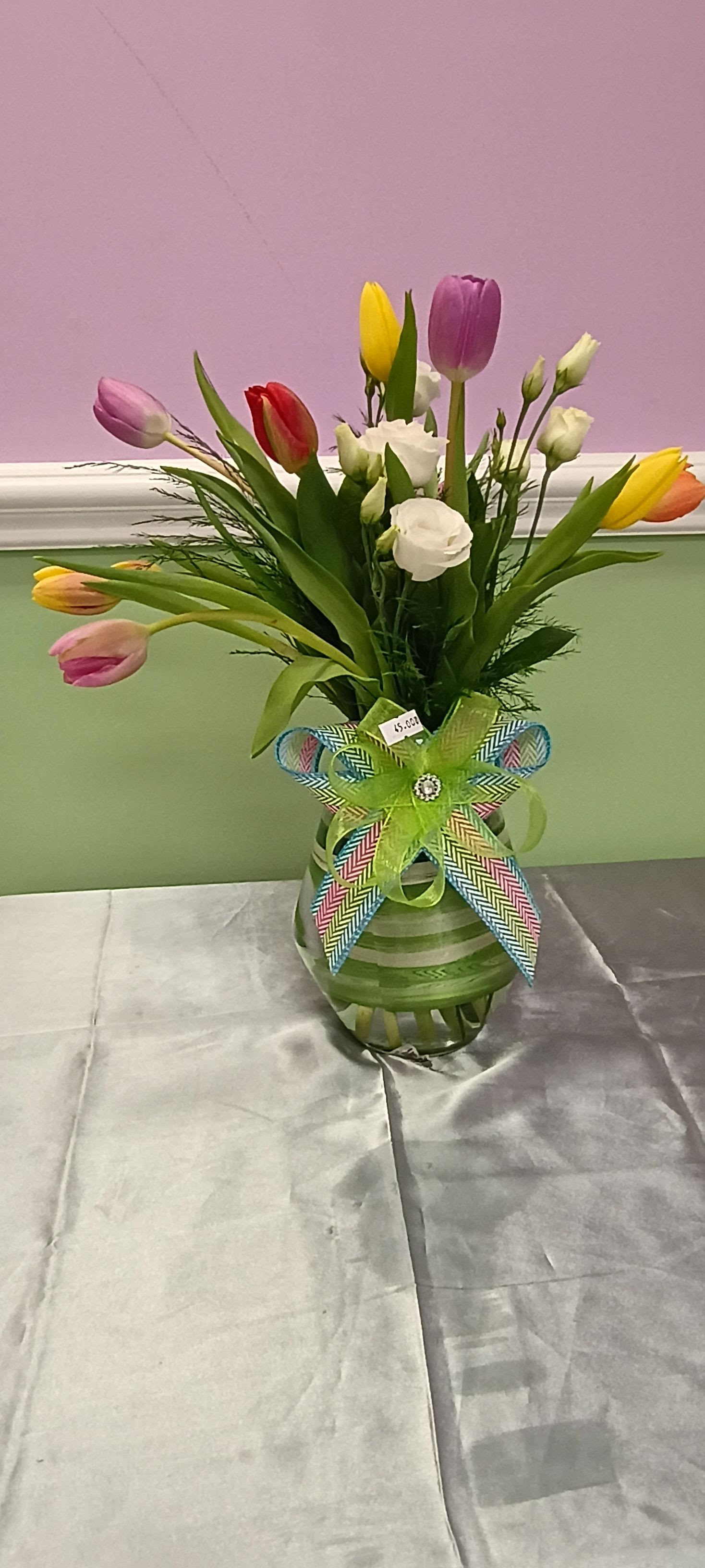 Elegant mixed color tulip arrangement with spray rose. - A cheerful cluster of mixed-color tulips creates an immediate springtime lift-perfect as a birthday bouquet, seasonal gift, or everyday celebration. This tulip arrangement with spray rose accents reads bright and joyful in pink, purple, yellow, red, and white tones, making the recipient feel genuinely seen and celebrated with a casual, hand-arranged charm.  Loose, upright tulip stems are arranged in a clear glass vase wrapped in green-striped material and finished with a large multicolored ribbon bow and a price tag. The composition feels informal and fresh, with varied heights and soft, natural movement that highlights the tulips' smooth petals and lively color contrasts. The vase and ribbon are included for an easy, ready-to-display gift.  Ideal for mothers, friends, coworkers, or anyone who loves cheerful spring color, this arrangement arrives ready to brighten a kitchen table, office desk, or entryway. Change the water every couple of days, trim stems at an angle before placing them back in fresh water, and keep away from direct heat to help blooms last longer.