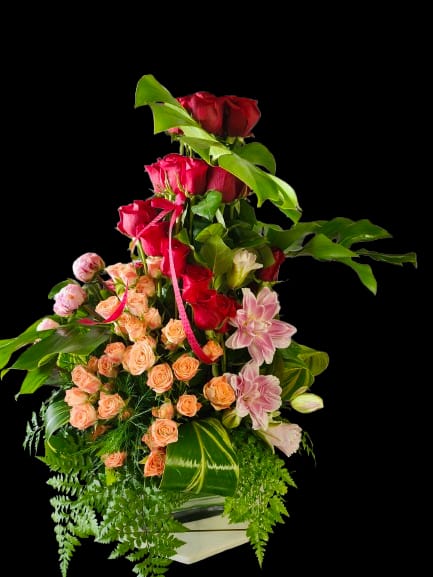 Muted Desire - Deep red roses stacked at the peak give this tall flower arrangement a dramatic, romantic silhouette-perfect as an anniversary roses display or a birthday bouquet for someone special. The vertical composition and lush tropical foliage make a bold statement that reads formal and celebratory, so the person you're thinking of will feel truly honored and seen.  Peach spray roses thread through the middle while pale pink lilies and buds soften one side, creating a warm gradient from crimson to blush. Broad tropical leaves and trailing fern fronds add texture and movement against the white container, which is finished with a pink ribbon for a polished gift-ready look. The arrangement's substantial height and layered design make it ideal for placing on an entry table, mantel, or beside a dining centerpiece for a memorable surprise.